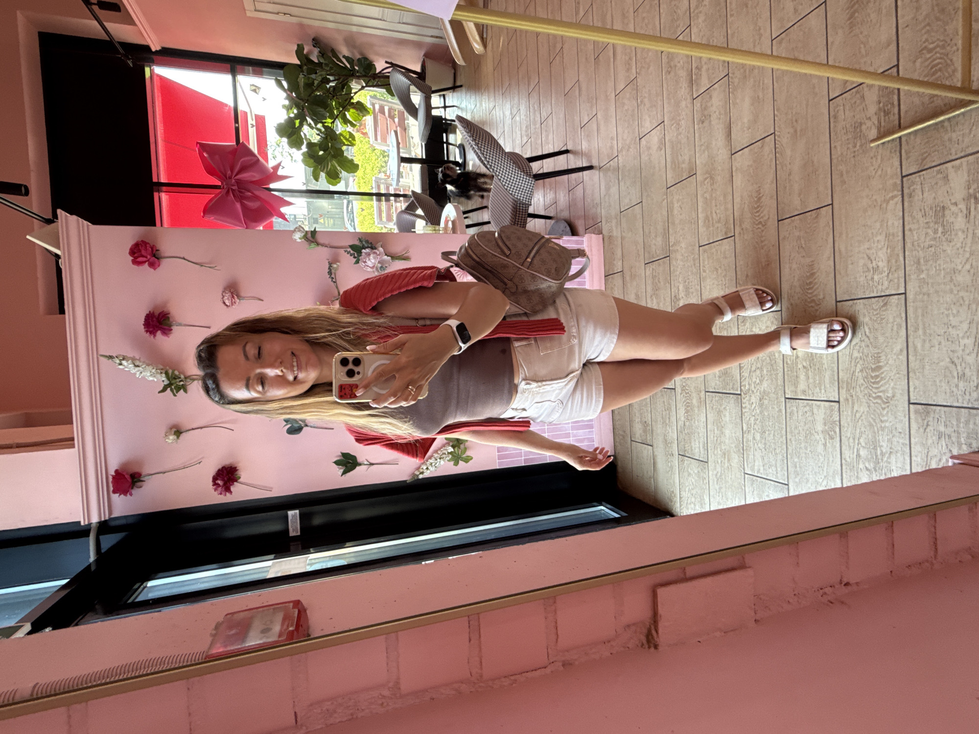 A woman stands in a brightly lit, pink-themed room, taking a mirror selfie. She is wearing white shorts, a grey tank top, and a red cardigan draped over her shoulders. A small, brown backpack is slung over her left shoulder. Her right hand holds a smartphone, and a white fitness tracker is visible on her left wrist. In the background, through a large window, an outdoor seating area with tables and chairs can be seen. A small black dog stands on the floor in the distance. The room is decorated with artificial pink and white flowers on a pale pink wall. A large pink bow is visible hanging near the window. The floor is tiled with a wood-like pattern.
