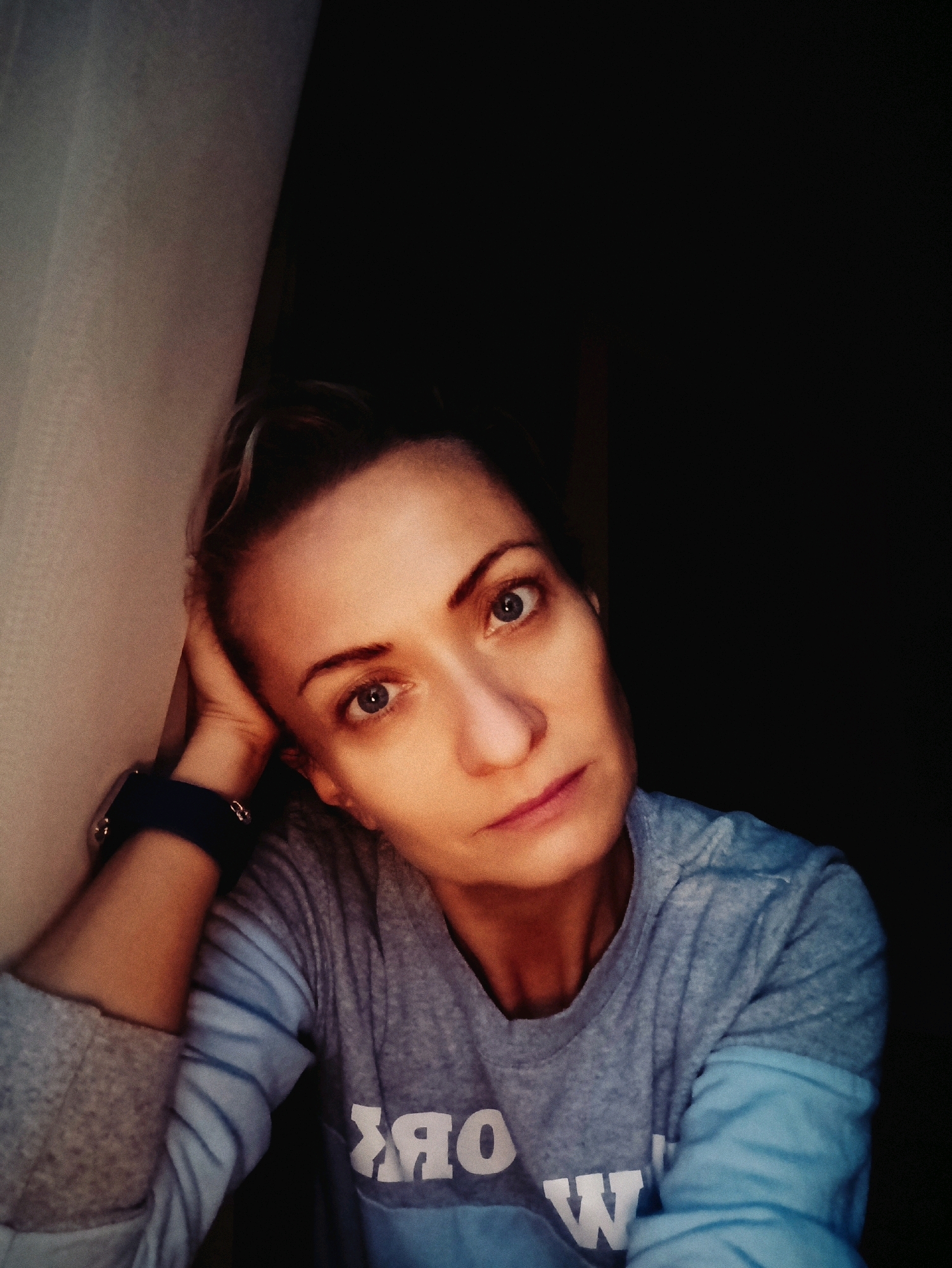 A selfie of a woman with blonde hair pulled back is taken indoors, with her left arm propping her head. She is wearing a blue and gray long-sleeved shirt. The background is divided between a light-colored textured surface on the left and a dark, indistinct background on the right. The lighting suggests artificial illumination, casting shadows on her face. The image does not provide any specific details about the location in Moscow, Russia, or any discernible forensic information.