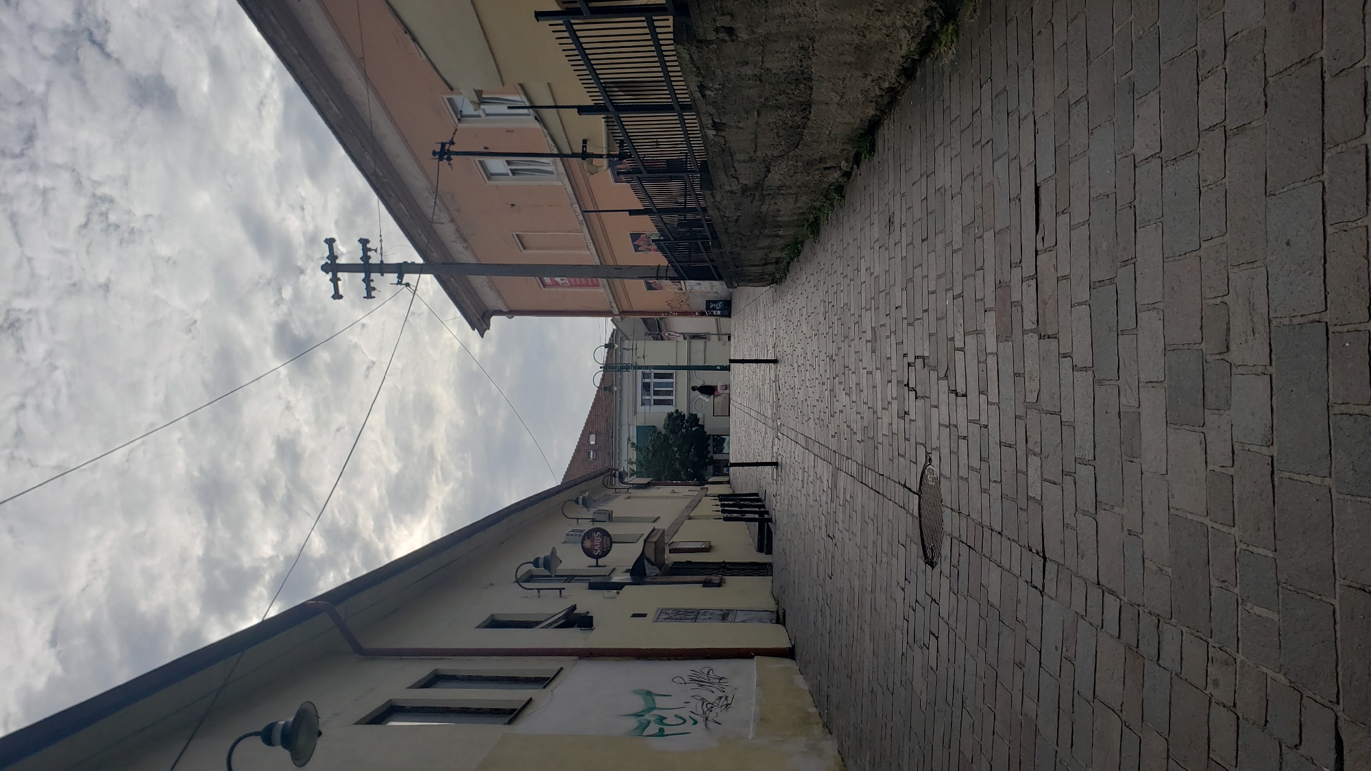 The image depicts an uphill cobblestone street in the historic center of Prešov, Slovakia. The street is lined with buildings of varying architectural styles and colors. On the left, a building with a yellow facade features graffiti and a sign for "SARIS." A street lamp is visible on the upper left. On the right, a peach-colored building is bordered by a black metal fence atop a stone retaining wall. A lone figure, dressed in dark clothing, is walking uphill in the distance, carrying a light-colored bag. Overhead, power lines crisscross against a cloudy sky. The cobblestone paving shows signs of wear and age, with some stones unevenly placed or cracked. The overall impression is one of a quiet, possibly older, urban street scene.