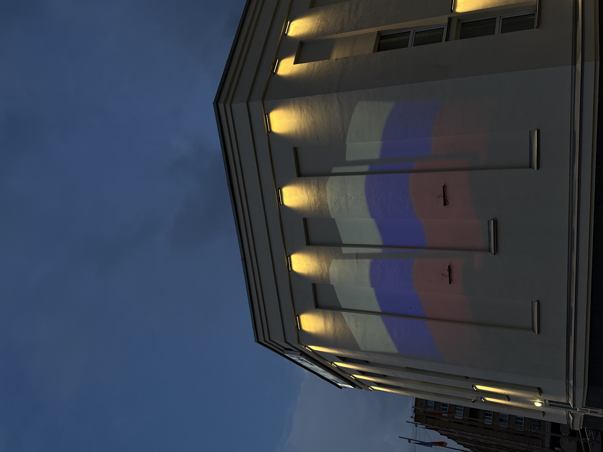 The image shows a building with the Russian flag projected onto its facade at dusk. The building is a light color and has several rectangular lights illuminating the facade from above. The Russian flag is projected in large sections of red, white, and blue, covering a significant portion of the building's wall. In the background, another building is visible, along with flags on poles. The sky is a dark blue, indicating it is evening. The scene appears to be outdoors in an urban environment.
