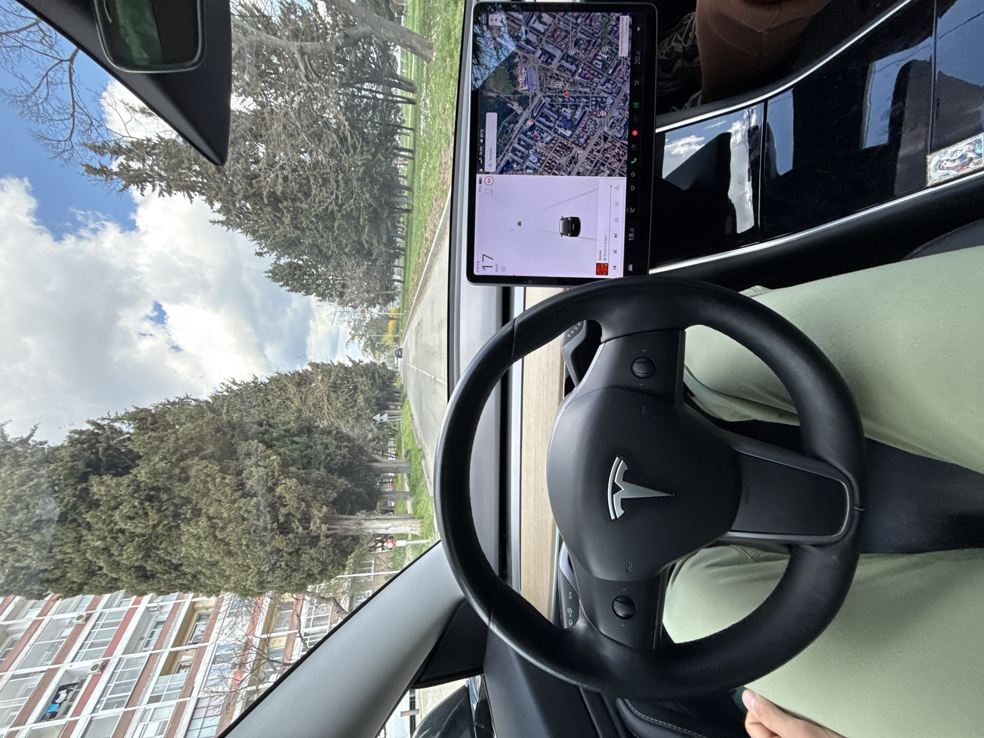 The image shows the interior of a Tesla car, specifically focusing on the steering wheel and the dashboard screen. The screen displays a navigation map, suggesting the car is in motion. The driver is wearing light green pants. Through the windshield, there is a view of a road lined with trees, and buildings are visible in the background. Based on the street layout and building architecture visible through the front window, the location is likely the city of Stara Zagora, Bulgaria. The sky is partly cloudy, indicating daytime.