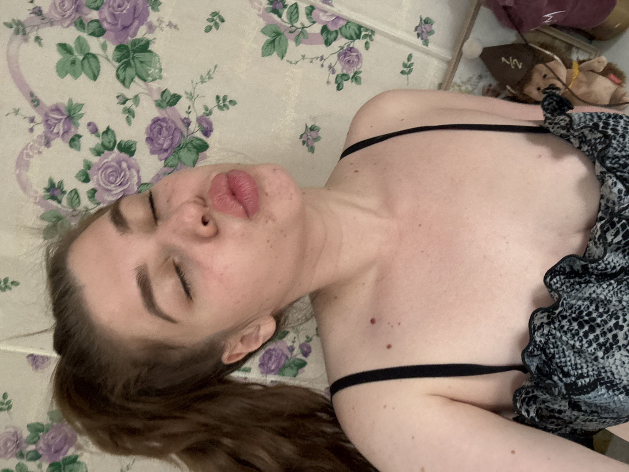 A woman is lying on a surface with a floral patterned covering. She has fair skin, brown hair pulled back in a ponytail, and is wearing a black top with thin straps. Her eyes are closed, and her lips are slightly puckered. A patterned gray and black fabric is draped over her chest. In the background, there is a stuffed toy with a hat. There is no information available to determine the location in the city of Мещёры, Russia.