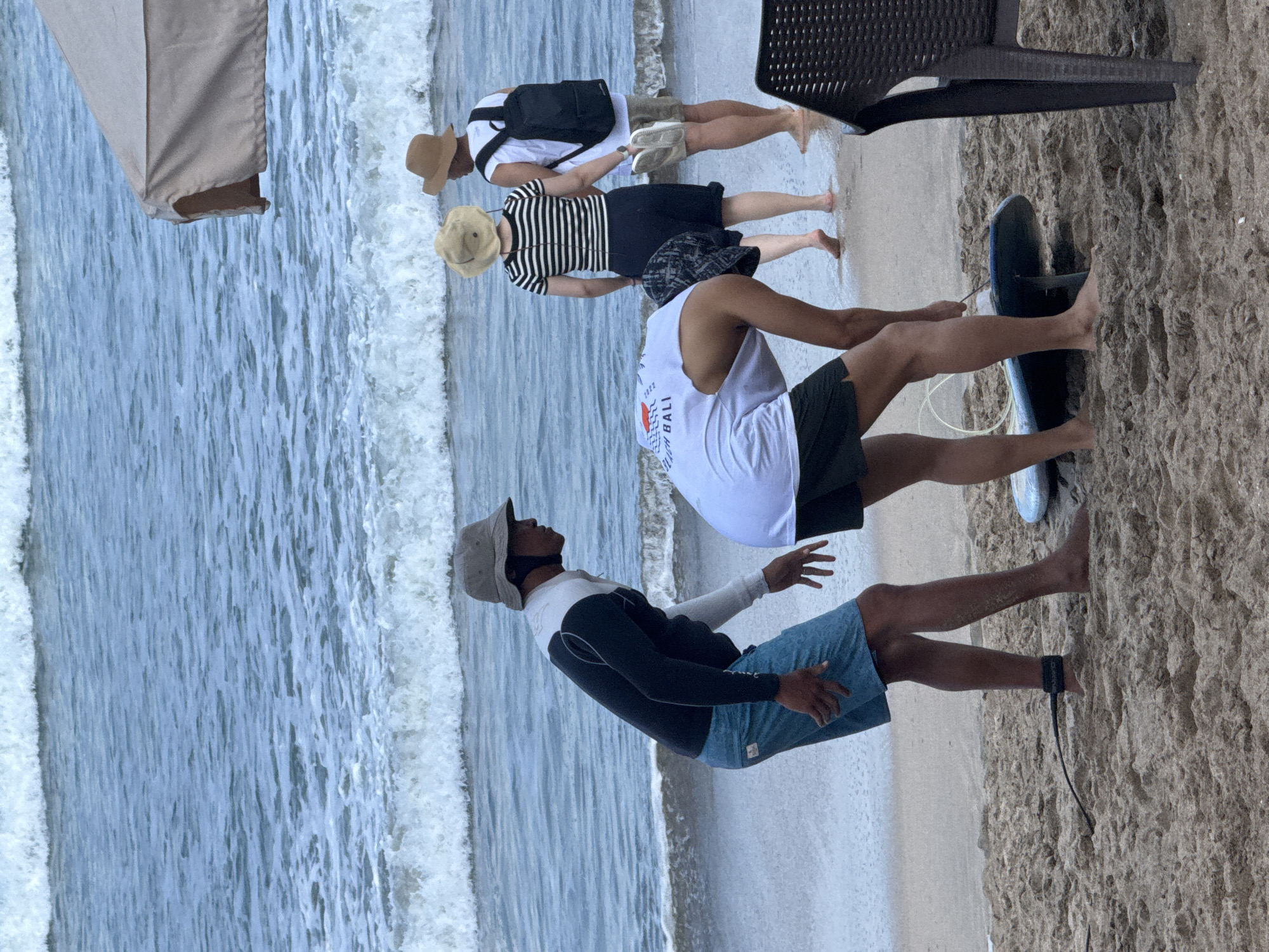 A group of four people are on the sandy shore of Kuta Beach, Indonesia, with the ocean and waves in the background. In the foreground, two individuals are interacting with a surfboard. One person, wearing a gray bucket hat, a black and white long-sleeved shirt, and blue patterned shorts, is bending over and appears to be adjusting something on the surfboard. A black leash is attached to their ankle. Another person, wearing a white t-shirt and dark shorts, is also bending over the surfboard, their back to the camera. Further back, two more individuals are standing near the water. One person wears a straw hat, a black and white striped shirt, and a dark skirt, holding a dark object in their hand. The other person, also wearing a straw hat, has a white t-shirt, khaki shorts, and a black backpack, and is holding a pair of white flip-flops. A tan umbrella canopy is visible at the top right of the frame, and a black chair is in the lower right corner. The sand is light brown and textured with footprints and some debris. The ocean water is blue-green with white waves breaking near the shore.