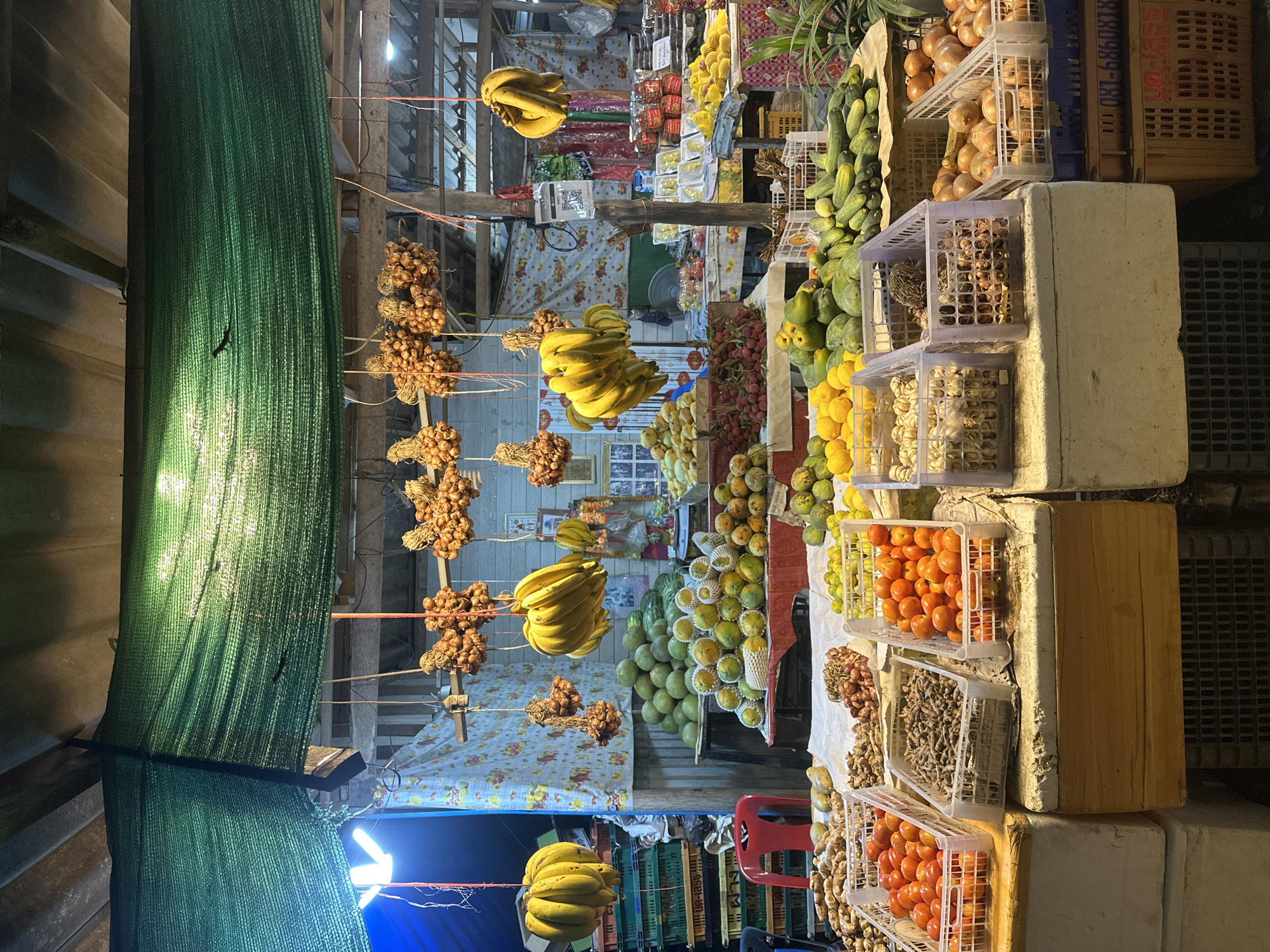 The image depicts a vibrant market stall in Koh Phangan, Thailand, likely in the evening or at night due to the artificial lighting. The stall is overflowing with a colorful assortment of fresh produce, including bananas, tomatoes, onions, mangoes, and other fruits and vegetables. The items are displayed in various containers, such as plastic crates and woven baskets, with some produce, like bunches of bananas and onions, hanging from strings. A green tarp or net is draped on the left side of the stall, providing shade or enclosure. In the background, glimpses of wooden structures and more crates suggest a bustling market environment. The scene conveys a sense of abundance and a lively atmosphere typical of a Thai market. There are no people visible in the image, and no discernible weather conditions can be inferred.