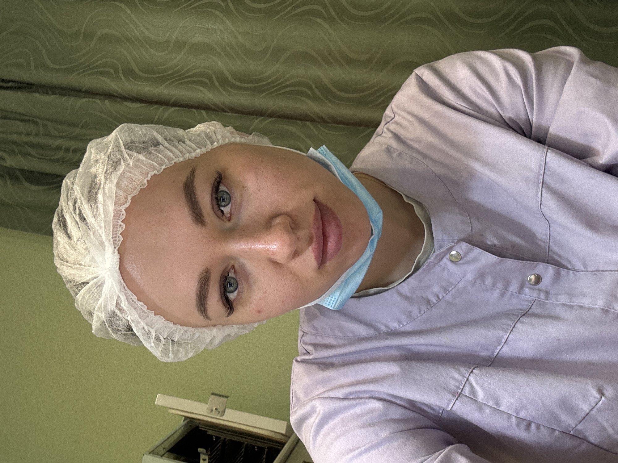 A woman wearing a white surgical cap and a light purple button-down medical scrub shirt is looking into the camera. She has a blue surgical mask pulled down below her chin. The background consists of a green wall with a wavy pattern at the top, and a white cabinet with black features at the bottom. The location is Ферма, Russia.