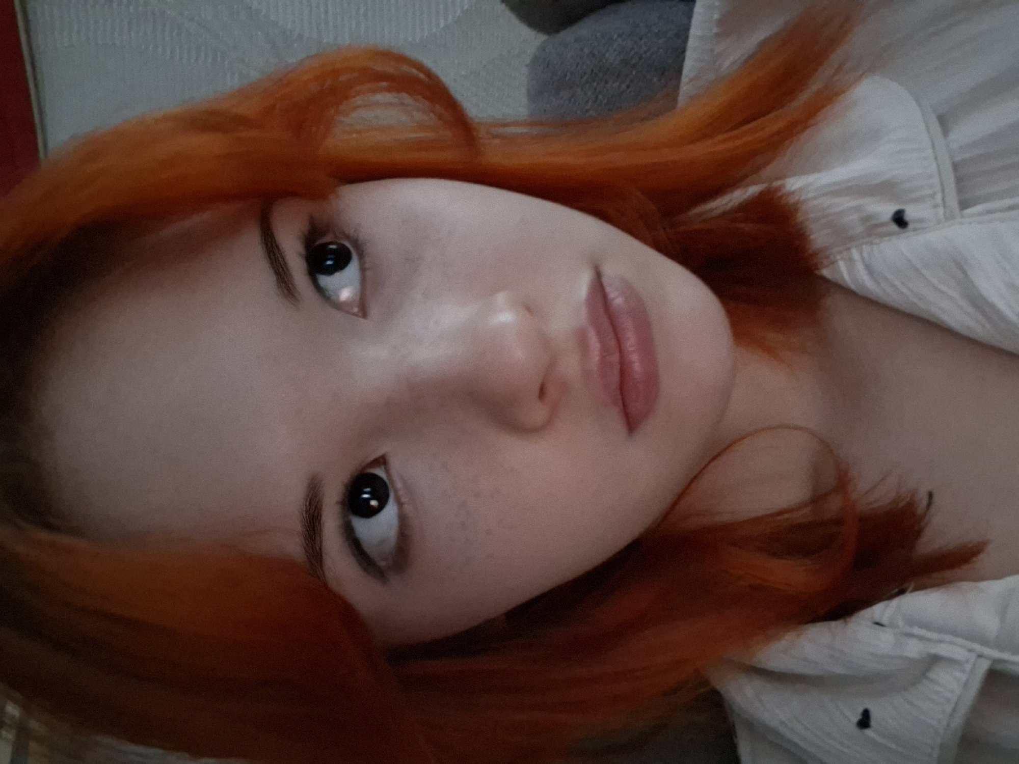 A close-up, high-angle portrait features a young person with vibrant red-orange hair, lying down or leaning back indoors in Chaikovsky, Russia. The individual has fair skin with subtle freckles visible on the nose and cheek. Their dark eyes are wide open, looking directly upwards, possibly at the camera. The lips are lightly parted, showing a natural pink hue, and appear full.
The lighting is soft, highlighting the contours of the face and creating a gentle sheen on the skin, particularly around the nose and lips. The person's hair frames their face and extends towards the top and bottom of the frame. They are wearing a light-colored, possibly white or off-white, collared shirt with some texture and visible dark buttons.
The background behind the head is a mix of a light, textured surface, possibly a wall or upholstered furniture, and a soft, grey fabric, suggesting an indoor, casual setting. The overall mood is calm and contemplative. No text is visible in the image.