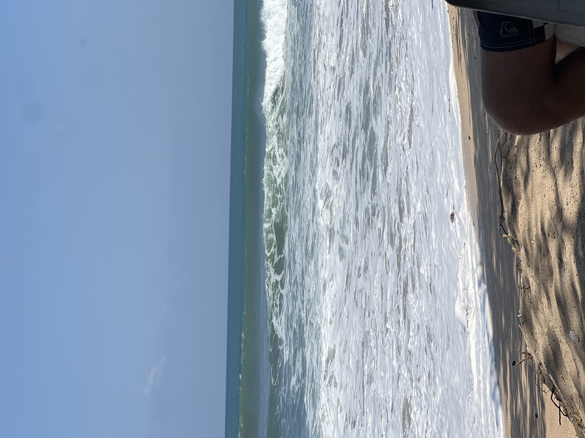 The image shows a beach scene in Kuta, Indonesia. The ocean is calm with gentle waves rolling onto the shore. The sky is clear and blue. In the foreground, a person is sitting on the sand, wearing swim trunks. The sand is light brown and there are some shadows cast by trees. The overall impression is one of a peaceful day at the beach.