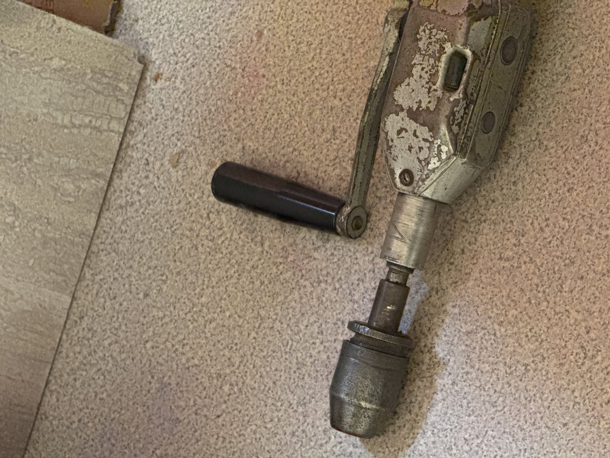 The image shows a close-up, high-angle shot of an old, manual hand drill resting on a speckled, textured surface that appears to be linoleum flooring. The drill is made of metal with a worn, chipped silver paint finish. A black handle is attached to a crank mechanism on the side of the drill. The drill's chuck, a cylindrical metal component used to grip drill bits, is visible at the bottom of the image. The background consists of the speckled floor and a section of what looks like a linoleum tile with a marbled pattern, positioned in the upper left corner. The lighting suggests an indoor setting, possibly a workshop or storage area. There is no visible text in the image. The overall impression is one of a vintage tool, likely unused for some time given its condition. The location context of Naro-Fominsk, Russia, does not provide any direct visual cues within the image itself.
