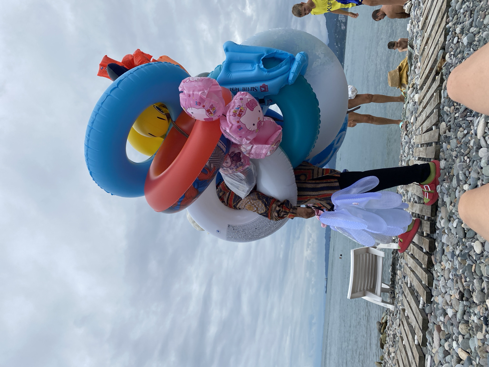On a pebble beach with wooden plank pathways and a white deck chair, a person in a patterned top and striped pants is carrying a large assortment of inflatable items, including a blue and white ring, a red and white ring, several pink Hello Kitty balloons, a yellow character balloon, and a blue inflatable swim vest. A person's legs in the foreground are visible. In the background, the Black Sea is calm under a cloudy sky. Several people are visible in the water and on the beach, including a boy in a yellow shirt with "KF" visible on it, a man, and a young child.