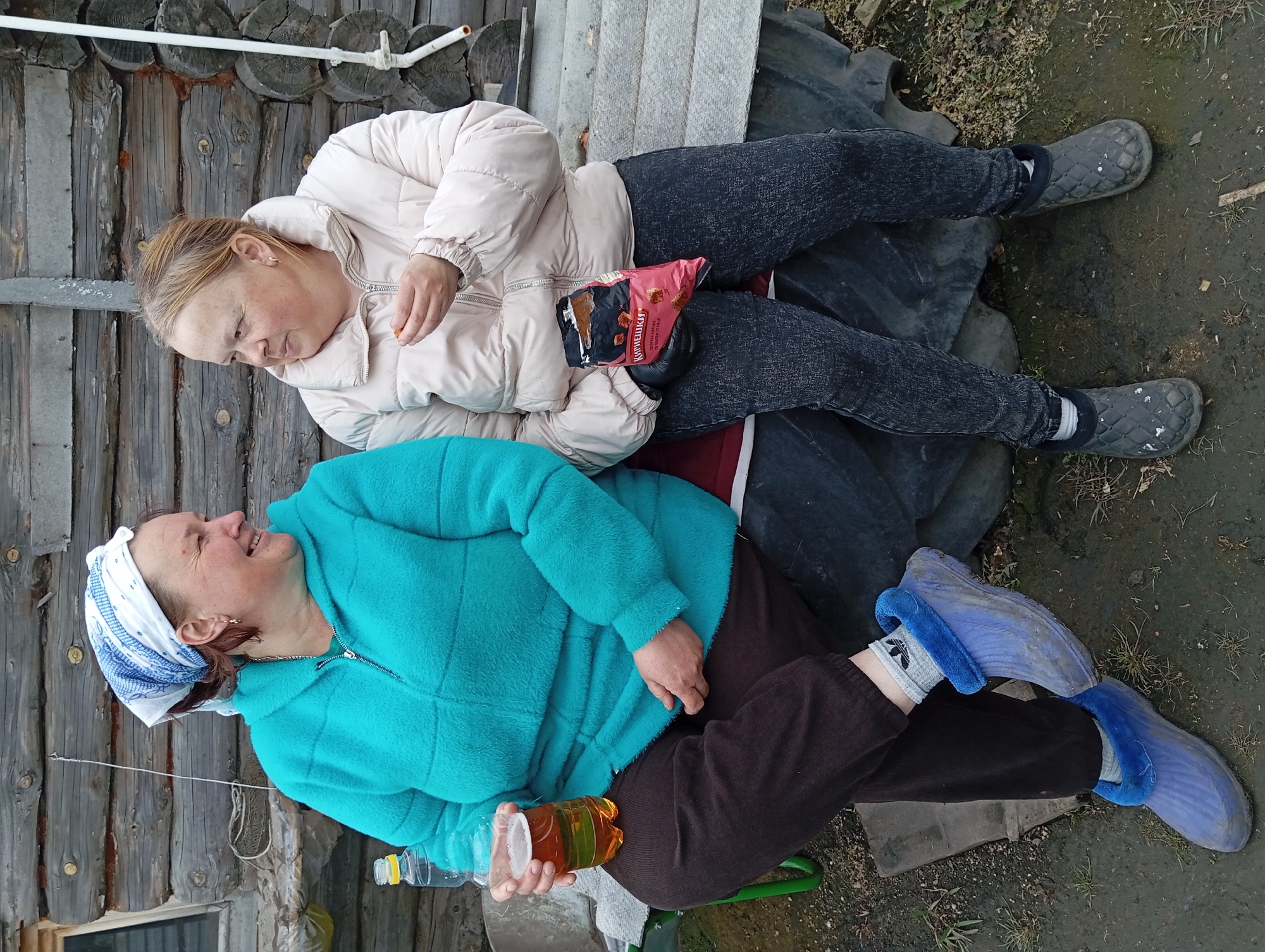 Two women are sitting outside a log cabin in Yurgamysh, Russia. One woman is wearing a white and blue headscarf, a turquoise jacket, and brown pants. She is holding a plastic cup with a golden liquid in it. The other woman is wearing a light pink jacket and dark jeans. She is eating from a pink bag of food. Both women are sitting on a dark-colored surface. The ground is muddy and there is some vegetation around them.