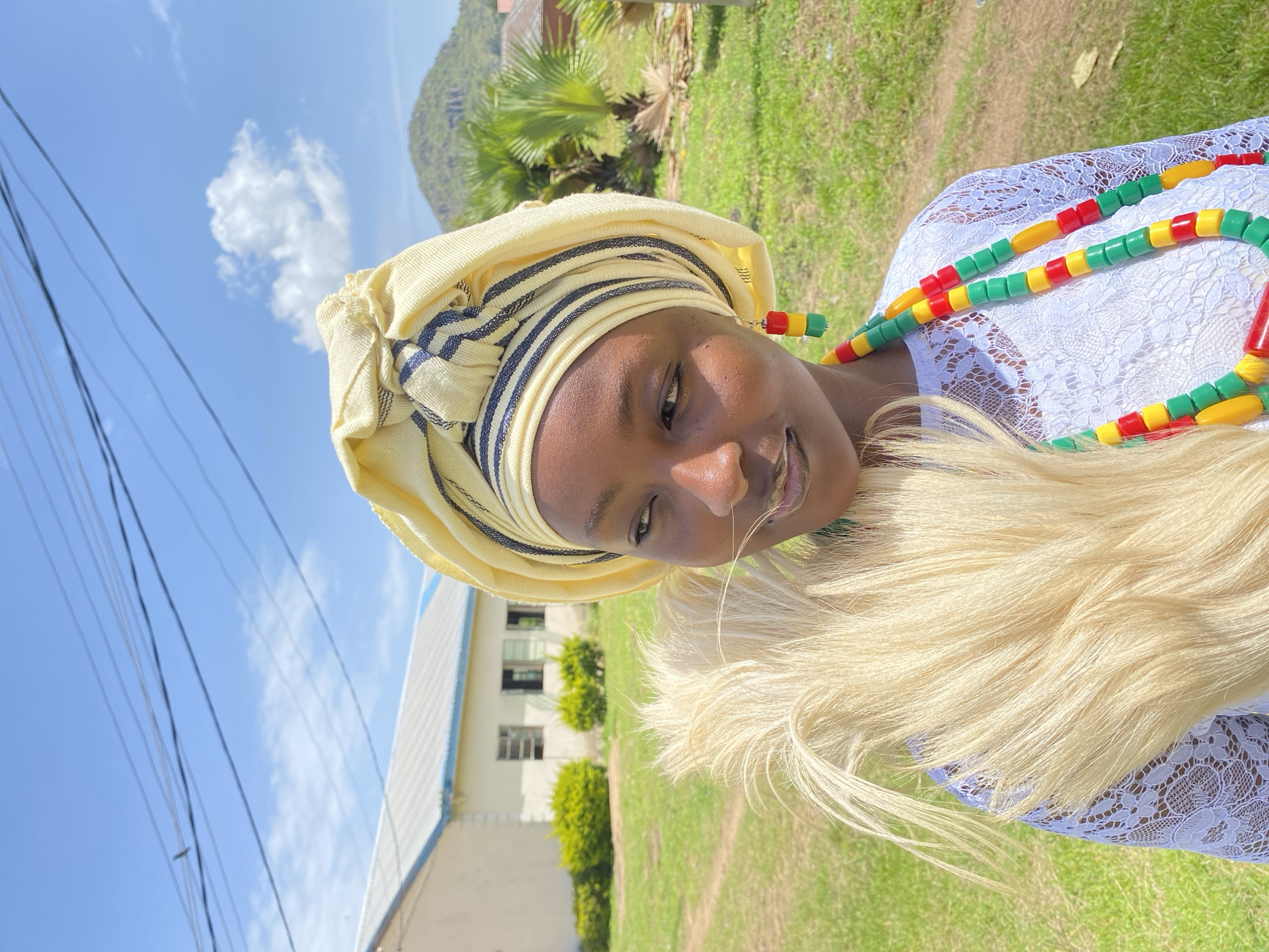 The media file is a portrait of a young Black woman wearing traditional attire, outdoors in Vom, Nigeria. She is adorned with a light yellow headscarf, accented with dark blue stripes. Around her neck, she wears a vibrant beaded necklace featuring red, green, and yellow beads, with a prominent faux fur stole draped around her shoulders. Her clothing appears to be a white, lace-like fabric.
The woman is looking towards the camera with a gentle smile, her right eye partly closed as if in a playful wink. The image is captured on a bright, sunny day, indicated by the clear blue sky with scattered white clouds and the green, well-maintained grassy surroundings. In the background, a light-colored building with visible windows and a blue-capped roof is present, along with some lush green bushes and trees, including a palm tree, characteristic of a tropical environment. Electrical wires are also visible in the sky, suggesting a populated area. The overall mood is cheerful and warm, capturing a moment of cultural expression and personal charm.