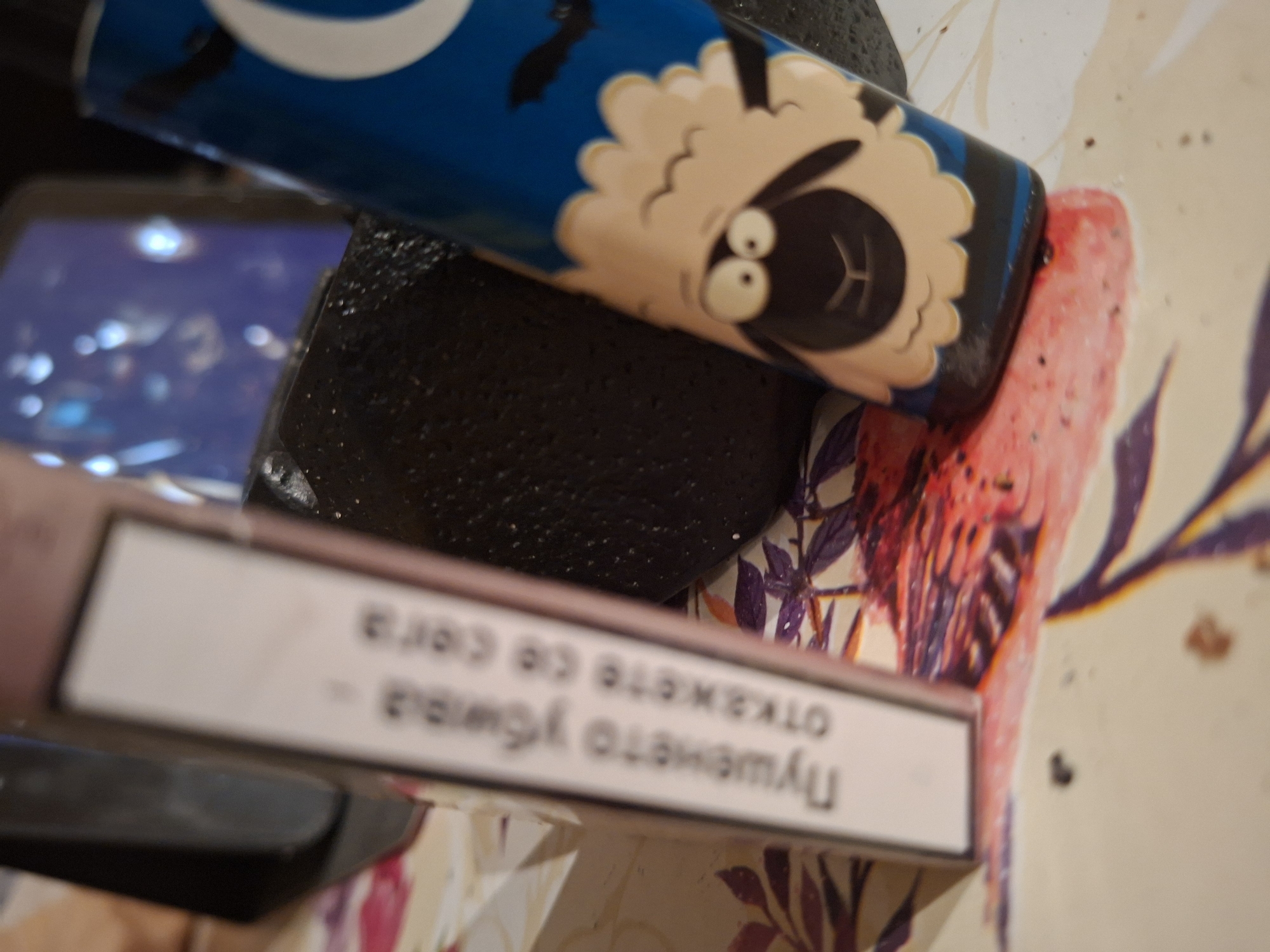 On a table with a floral pattern, there is a blue aerosol can with a cartoon sheep printed on it, a black lighter, a pink pack of cigarettes with text in Cyrillic characters, and a phone reflecting light. The location is likely in Plovdiv, Bulgaria given the language of the text on the cigarette pack.