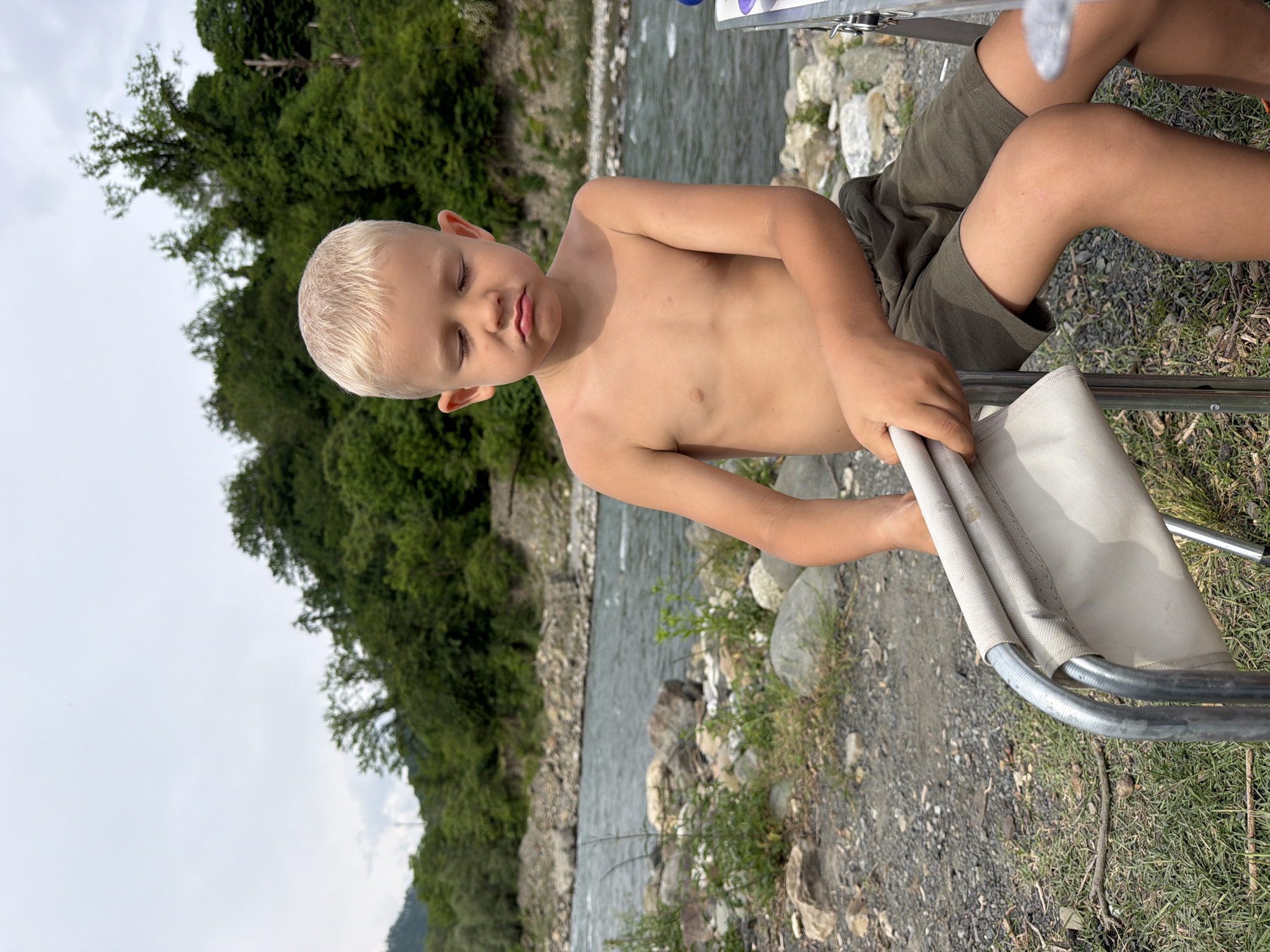 A young boy with blond hair is sitting outdoors, shirtless, with his eyes closed and his head tilted slightly. He is wearing olive-green shorts. He is sitting on a folding chair, the fabric of which is a light beige color. His right arm is resting on the chair, and his hand is holding onto the armrest. The setting appears to be near a river, with rocky terrain and dense green trees in the background. The sky is overcast. The overall impression is one of relaxation or a moment of quiet contemplation, possibly during a summer day. The location is identified as Sochi, Russia. There is no visible text in the image.