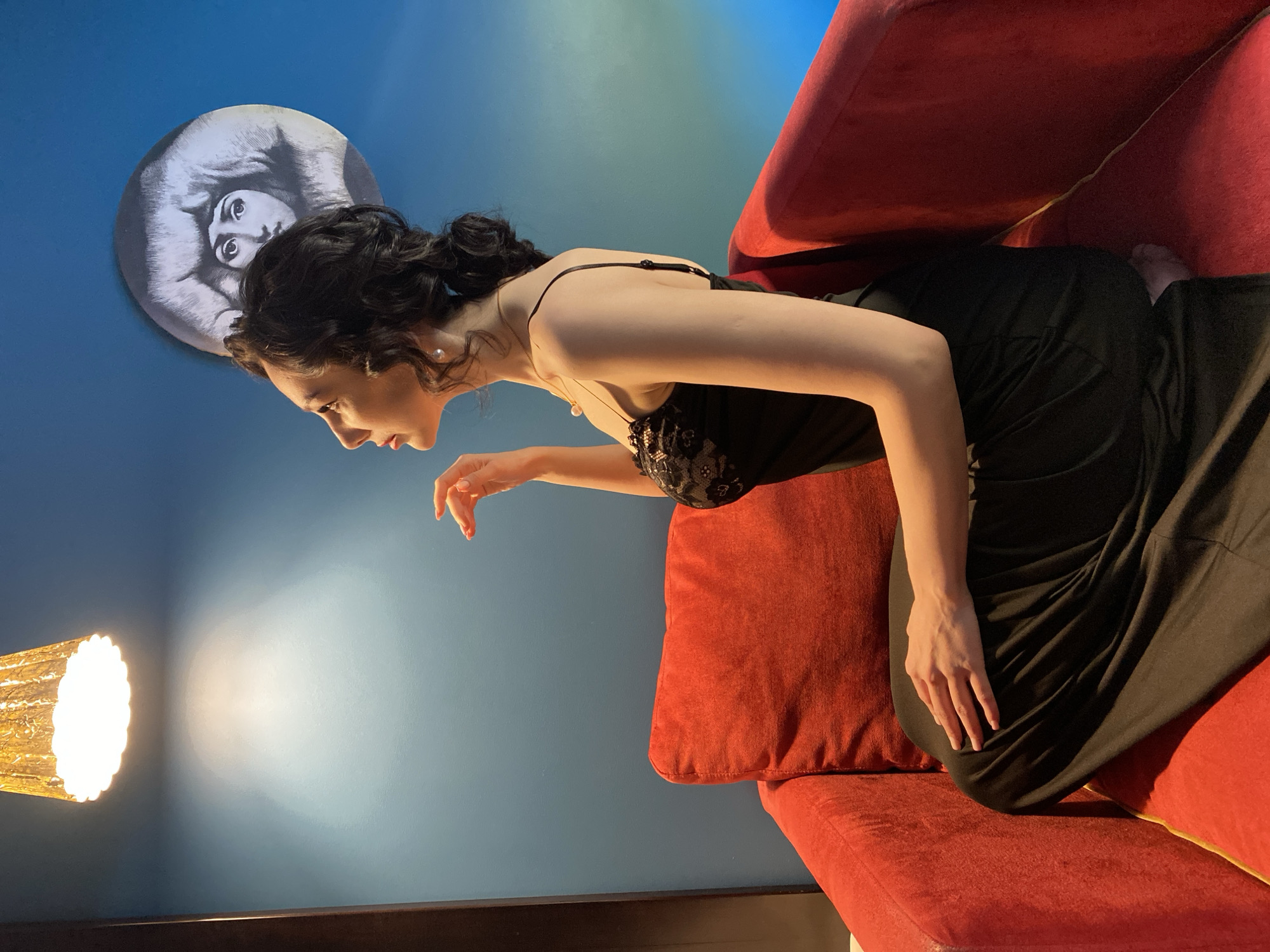 A woman in a black spaghetti strap dress sits on a red couch. Her dark hair is styled in an updo. She is leaning forward, looking downwards with her mouth slightly open, and her right hand is extended. The wall behind her is blue and has a circular art piece of a person's face on it. To the left, there is a lamp with a lit bulb. The room appears to be indoors. This photo could not be interpreted to be specifically within Yekaterinburg, Russia, without more information.