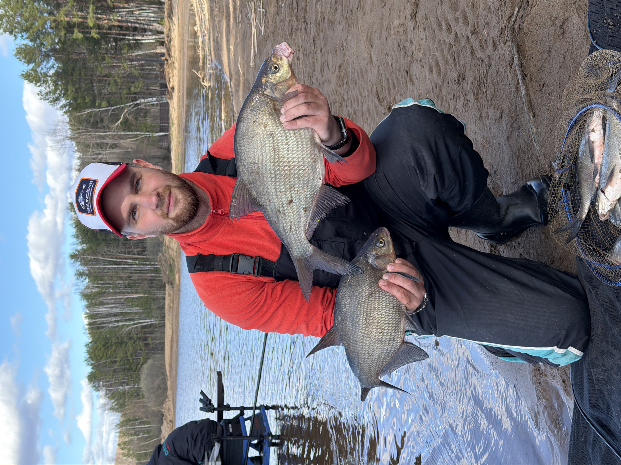 A man with a beard and a white and red cap is holding two fish. He is wearing a red jacket, black pants, and black boots. He is sitting on a sandy shore next to a body of water. There is a net full of fish to his right. The background shows trees and a blue sky with some clouds. The location is identified as the city of Chaikovsky, Russia.