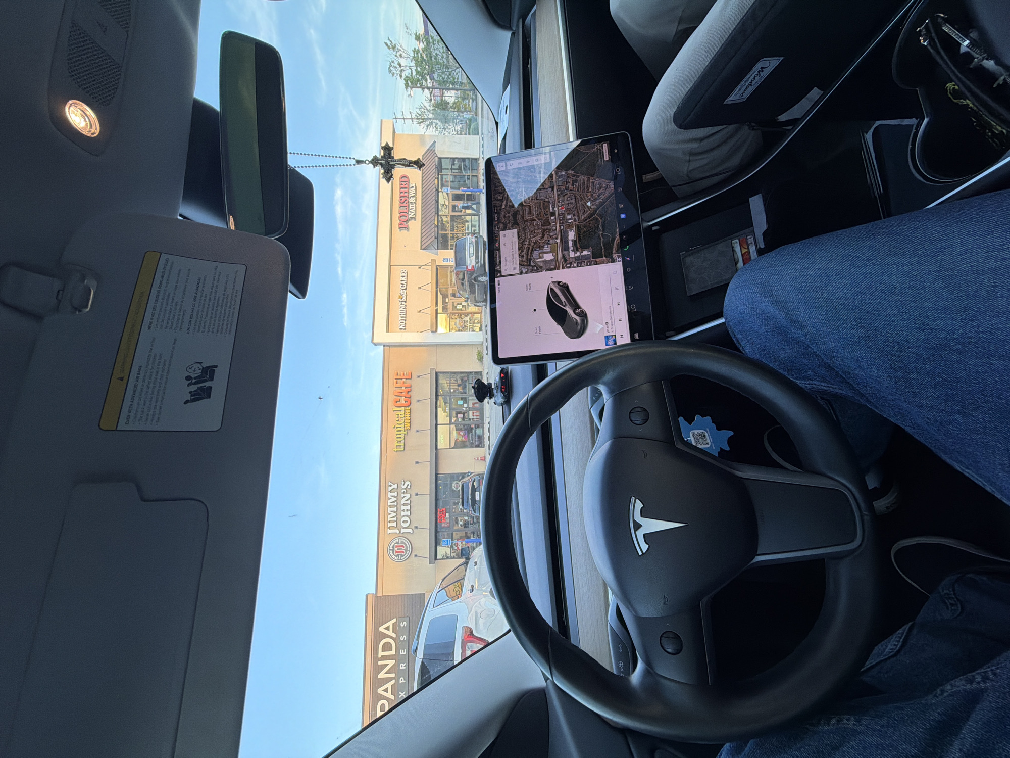 The image depicts the interior of a Tesla vehicle, specifically the dashboard and steering wheel. The Tesla logo is visible in the center of the steering wheel. The central display screen is on, showing a map and controls. The person in the driver seat wears blue jeans and black shoes.
Outside the car window, there is a view of a commercial area, featuring a storefront of PANDA XPRESS, Jimmy John's, Tropical CAFE, NOTHING CAKES and POLISHED NAIL & WAX. The buildings and businesses suggest that the location is in an urban or suburban area, potentially a shopping center or strip mall.
Based on the businesses listed, this location appears to be in Myrtle Beach, South Carolina.