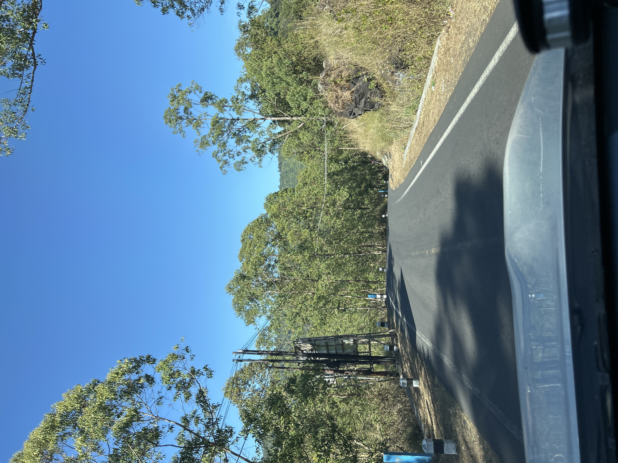 The image captures a view from a vehicle's dashboard looking forward on a paved road in Kintamani, Indonesia. The road curves gently to the right, bordered by dry grass, dense green trees, and a rocky outcrop on the right side. On the left, a utility pole with numerous wires stands beside the road, accompanied by several striped bollards. The sky is a clear, vibrant blue. The foreground of the image is dominated by the metallic and plastic interior of the vehicle's dashboard.