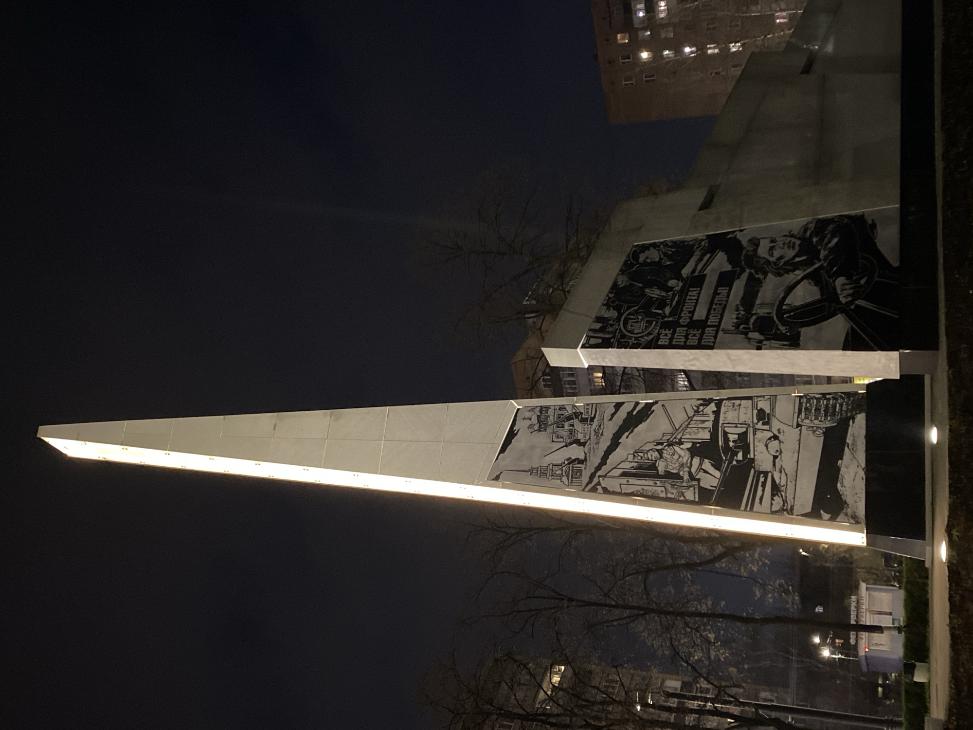 The image displays a towering, angular monument at night in Ryazan, Russia. The monument, constructed from a light-colored material, is brightly illuminated by an integrated strip light running along its main diagonal edge, casting a warm glow. The sky above is dark, indicative of nighttime, with distant artificial lights from buildings visible in the background.
The monument features multiple large murals or reliefs, rendered in a striking black and white graphic style. These panels depict scenes of human endeavor, particularly focusing on industrial work and wartime support. One prominent mural shows a figure, possibly a woman, engaged with machinery or a control panel, beneath the clear Cyrillic text: "ВСЁ ДЛЯ ФРОНТА! ВСЁ ДЛЯ ПОБЕДЫ!" (All for the Front! All for Victory!), a famous Soviet slogan from World War II. Other panels illustrate industrial machinery and workers. Additional visible Cyrillic text includes "ВСЁ ДЛЯ РОДИНЫ" (All for the Motherland) and "ЗА РОДИНУ" (For the Motherland), further emphasizing a patriotic and historical theme.
In the mid-ground and foreground, bare-branched trees are visible, silhouetted against the dark sky and partially illuminated by the monument's lights. A multi-story residential building with illuminated windows can be seen in the upper right background, confirming an urban setting. The monument's design and thematic murals suggest it serves as a commemorative tribute, likely honoring historical wartime efforts, labor, and patriotism.