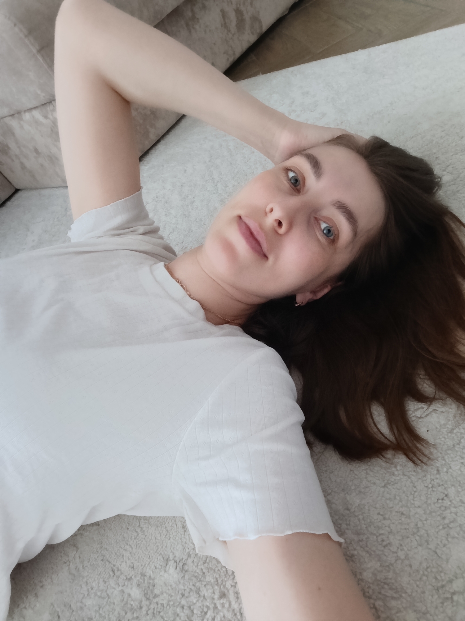 A female individual with dark brown hair and blue eyes is depicted lying on her back, looking directly at the camera. She is positioned on a light-colored, textured carpet. Her right arm is bent, with her hand resting behind her head, and her left arm is extended. She is wearing a white, short-sleeved top with a subtle textured pattern and a ruffled hem on the sleeves. A thin, gold-colored chain necklace is visible around her neck, and a small gold-colored hoop earring is present in her left ear. In the upper left background, a portion of a light-colored, upholstered sofa is visible. The specific city location of Chaikovsky, Russia, cannot be determined from the visual information provided in the image.