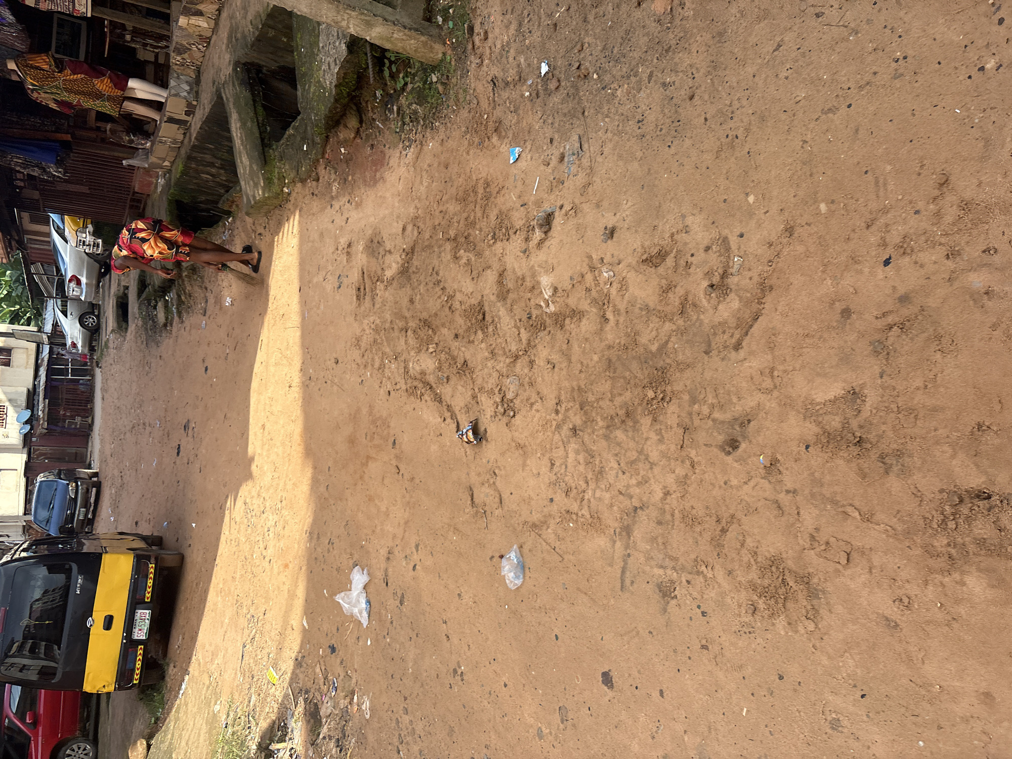 In Onitsha, Nigeria, a dirt road runs between buildings and open market stalls. A woman dressed in a vibrant floral dress is sweeping the ground with a broom. To her left, a yellow and black van is parked, its license plate reading "SSM 534 YB". Several cars are parked further down the road, and various debris, including plastic bags, is scattered across the ground. The area appears to be a bustling marketplace or residential street, typical of Onitsha's urban landscape.