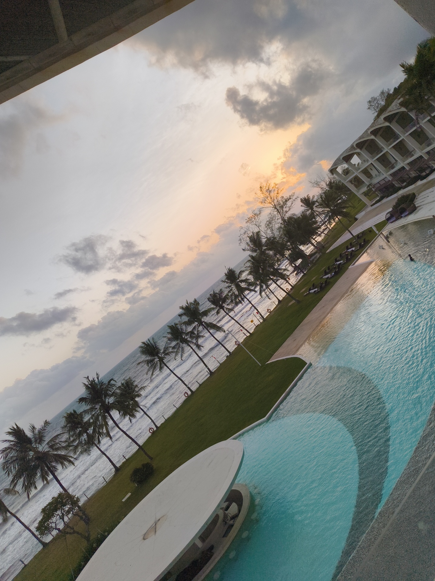This is an angled, outdoor, eye-level shot of a resort swimming pool and beach area at sunset in Phú Quốc, Vietnam. The scene is dominated by the large, multi-level swimming pool, which occupies the right side of the frame and features a gradient of blues, transitioning to a deeper shade near the right edge. A single person is visible swimming in the pool. Beyond the pool, a strip of green grass leads to a sandy beach lined with numerous palm trees. The ocean is visible behind the palm trees, with gentle waves breaking on the shore. In the distance, a multi-story building with a distinctive arched facade is partially visible on the right. The sky above is filled with a mix of gray clouds and areas of soft orange and yellow light, indicating the sunset. The overall impression is one of a serene tropical evening at a resort.