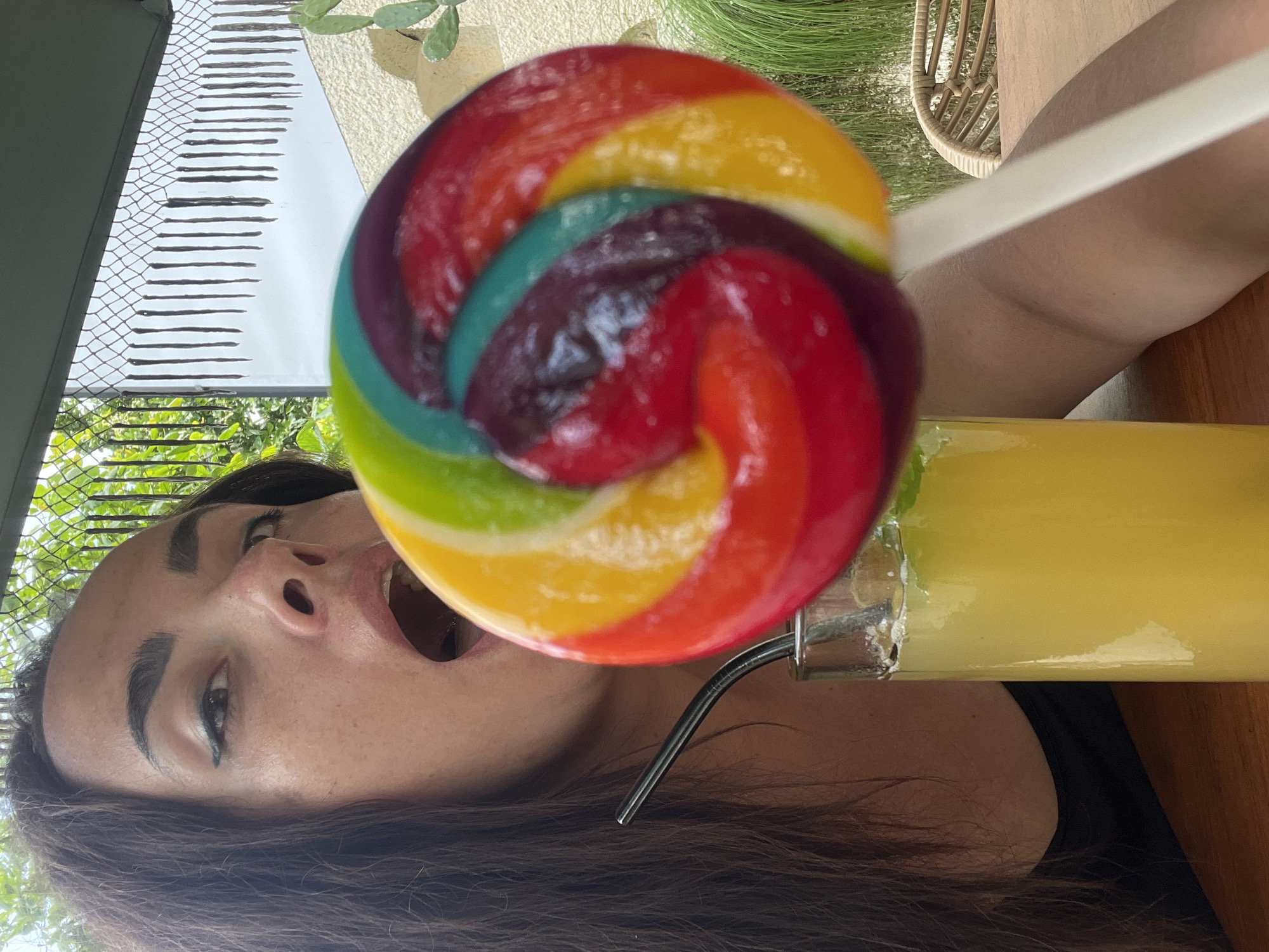 A woman, seen from the chest up, is holding a large, colorful lollipop in her right hand and a glass of yellow liquid with a metal straw in her left. The lollipop is positioned in front of her mouth, which is open. She is wearing a black top. The background shows an outdoor setting with a fence, greenery, and a beige wall. The scene appears to be taking place on a table. The woman's face is partially obscured by the lollipop.