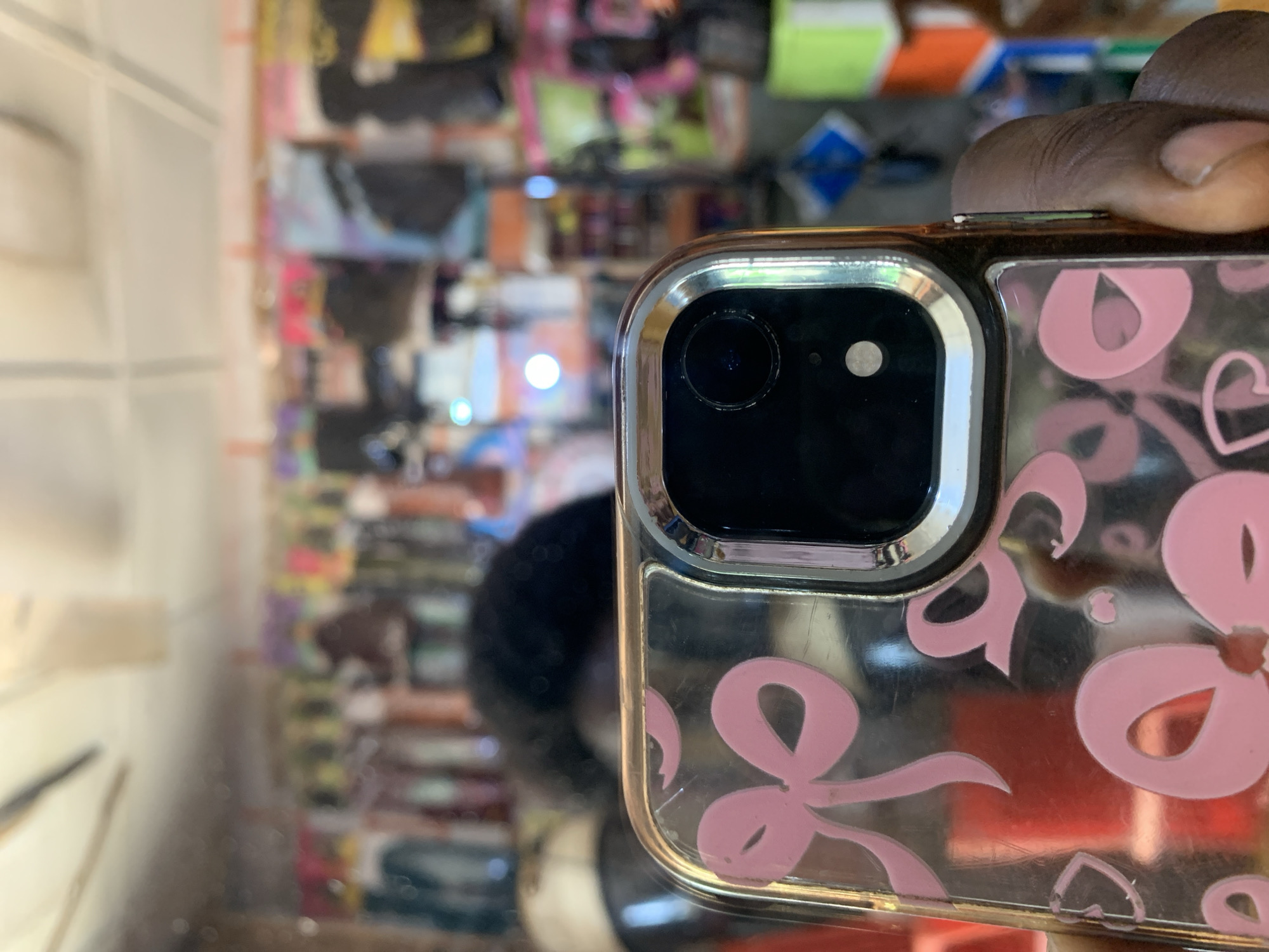 A close-up photograph shows a hand holding an iPhone with a clear case adorned with pink ribbons and hearts. The phone's camera module is visible. The background is blurry and appears to be a reflection of shelves stocked with various items. The location in the city of Rafin Par, Nigeria is not discernible from this image.