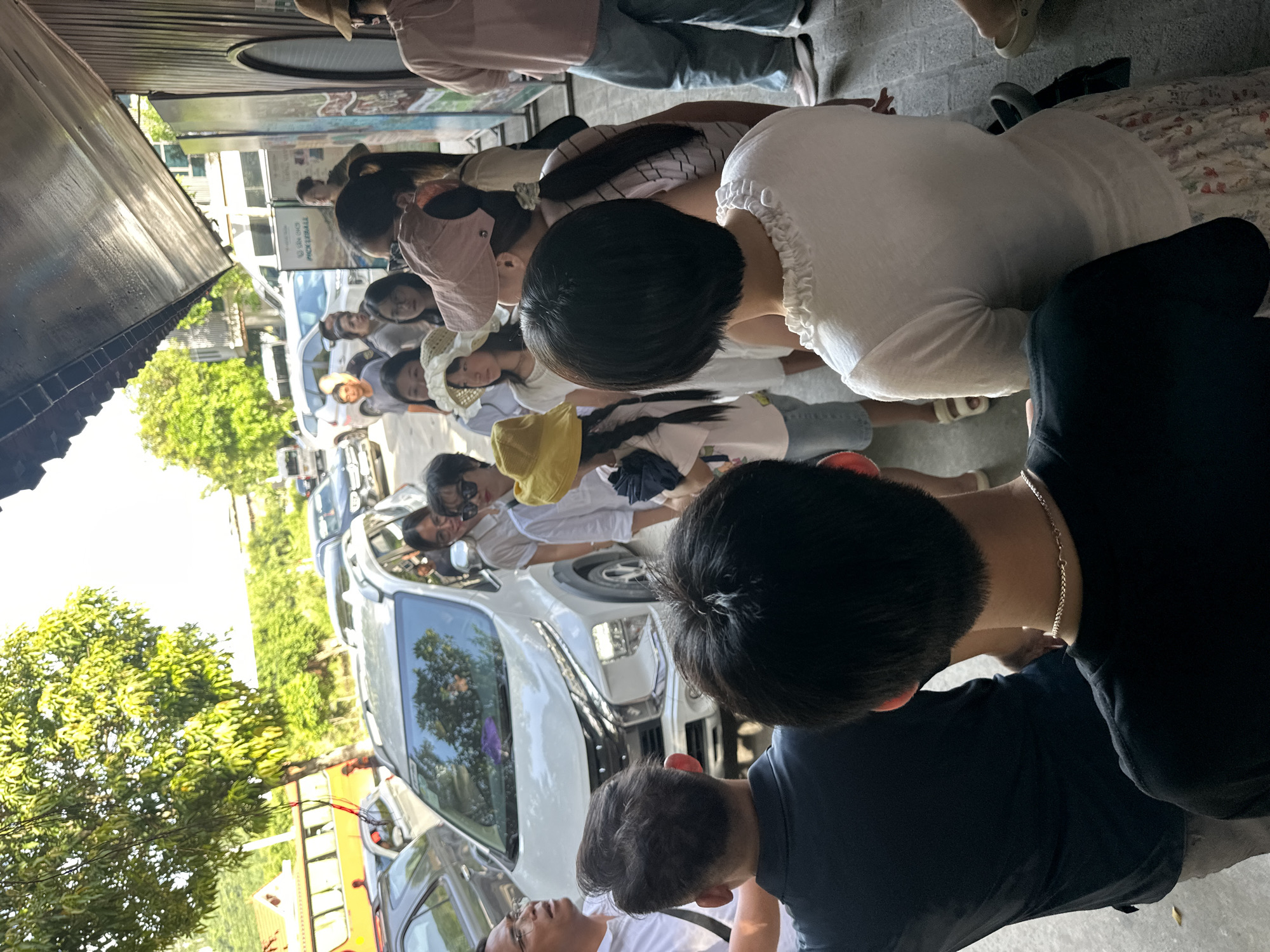 The image depicts a group of people gathered near a white SUV, possibly in a public area with a building entrance visible on the right. Several individuals are looking towards the camera or engaging in conversation. The environment appears to be an outdoor setting with trees and foliage visible in the background. The presence of a bus in the distance suggests a transportation hub or a tourist area. The scene seems to be captured during daylight hours, with shadows indicating the sun's position. The people are dressed in casual attire, suggesting a relaxed atmosphere.