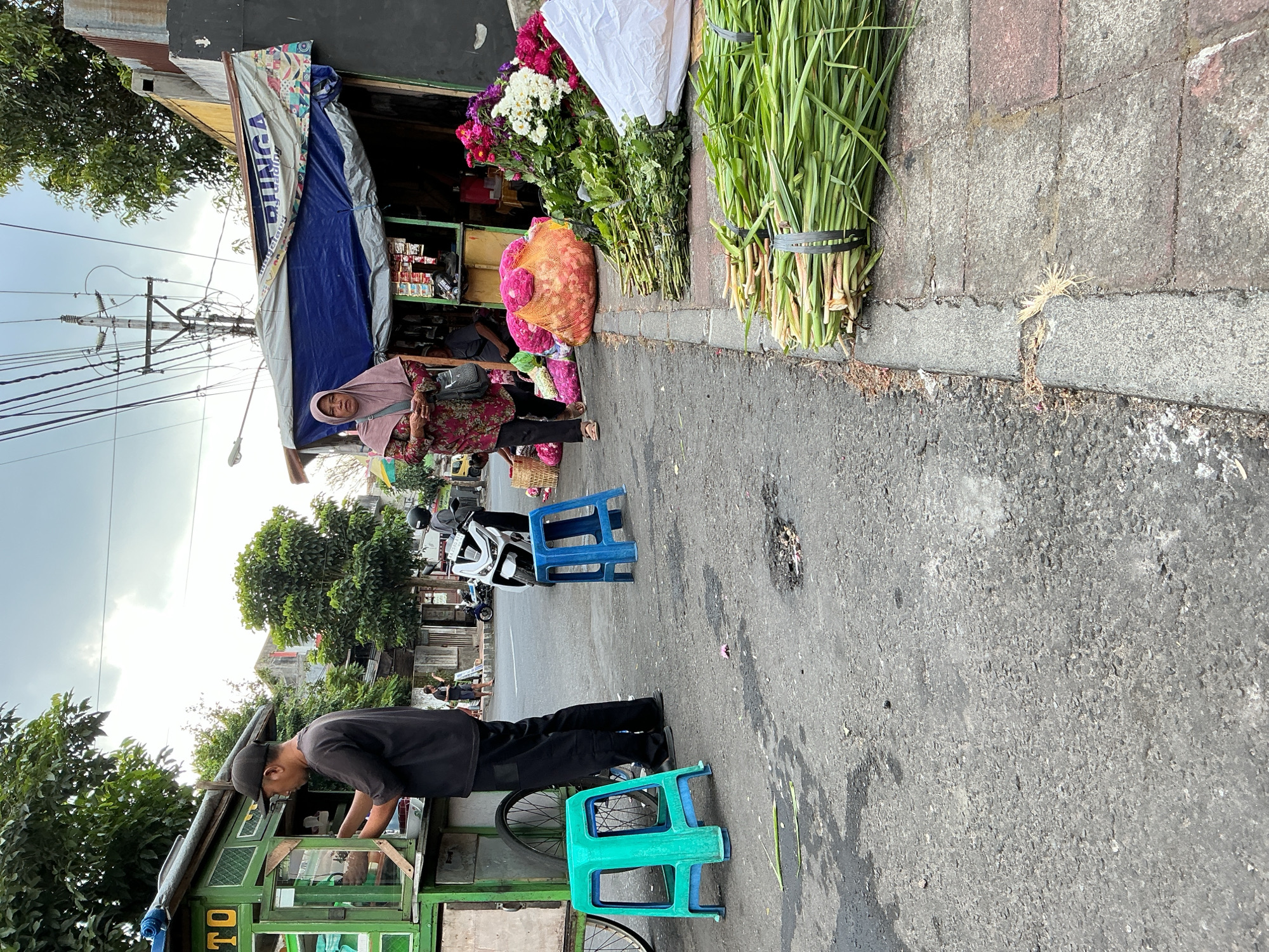 The image depicts a street scene in Magelang, Indonesia. A man wearing a black t-shirt and a black baseball cap is standing next to a green food cart, appearing to be serving a customer. To the right of the food cart, a woman in a pink headscarf and a patterned dress is walking. In the foreground, there are two plastic stools, one teal and one blue. The street is paved with asphalt and shows signs of wear and tear. On the right side of the street, there is a flower stall displaying various bundles of flowers and greenery. The stall is covered by a blue and white tarp. The background shows buildings, trees, and overhead power lines, indicative of an urban environment. A motorcycle is parked on the street near the flower stall. The overall scene suggests a lively street market or commercial area.