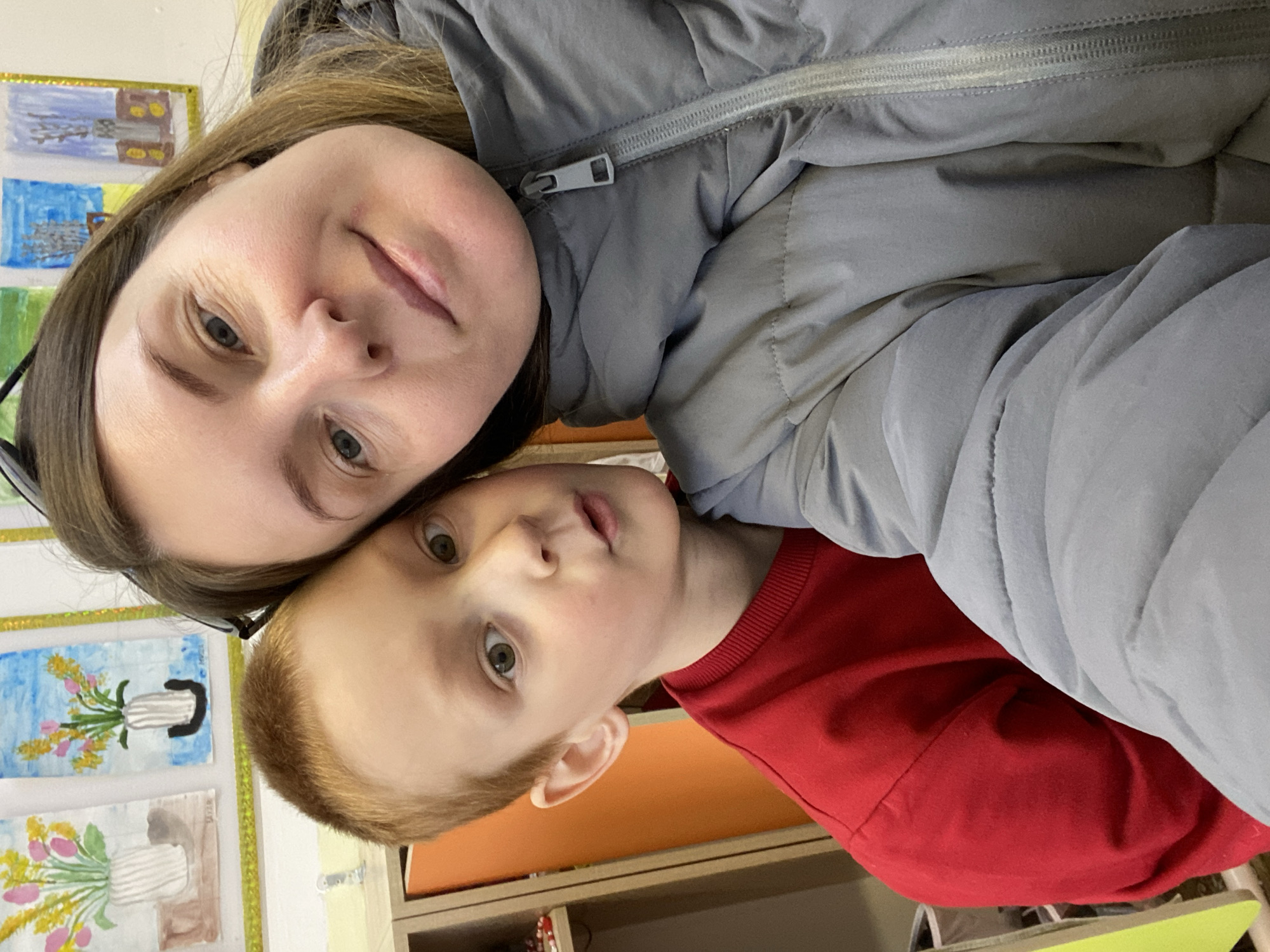 A woman and a young boy pose closely together for a selfie. The woman, on the upper left of the frame, has light skin, and brown hair. She is wearing a gray jacket with a zipper visible. The boy, on the lower right, has fair skin, light red hair, and is wearing a red long-sleeved shirt. They are indoors. Behind them are children's paintings in frames, a wooden shelf, and a green table. The location cannot be determined from the image.