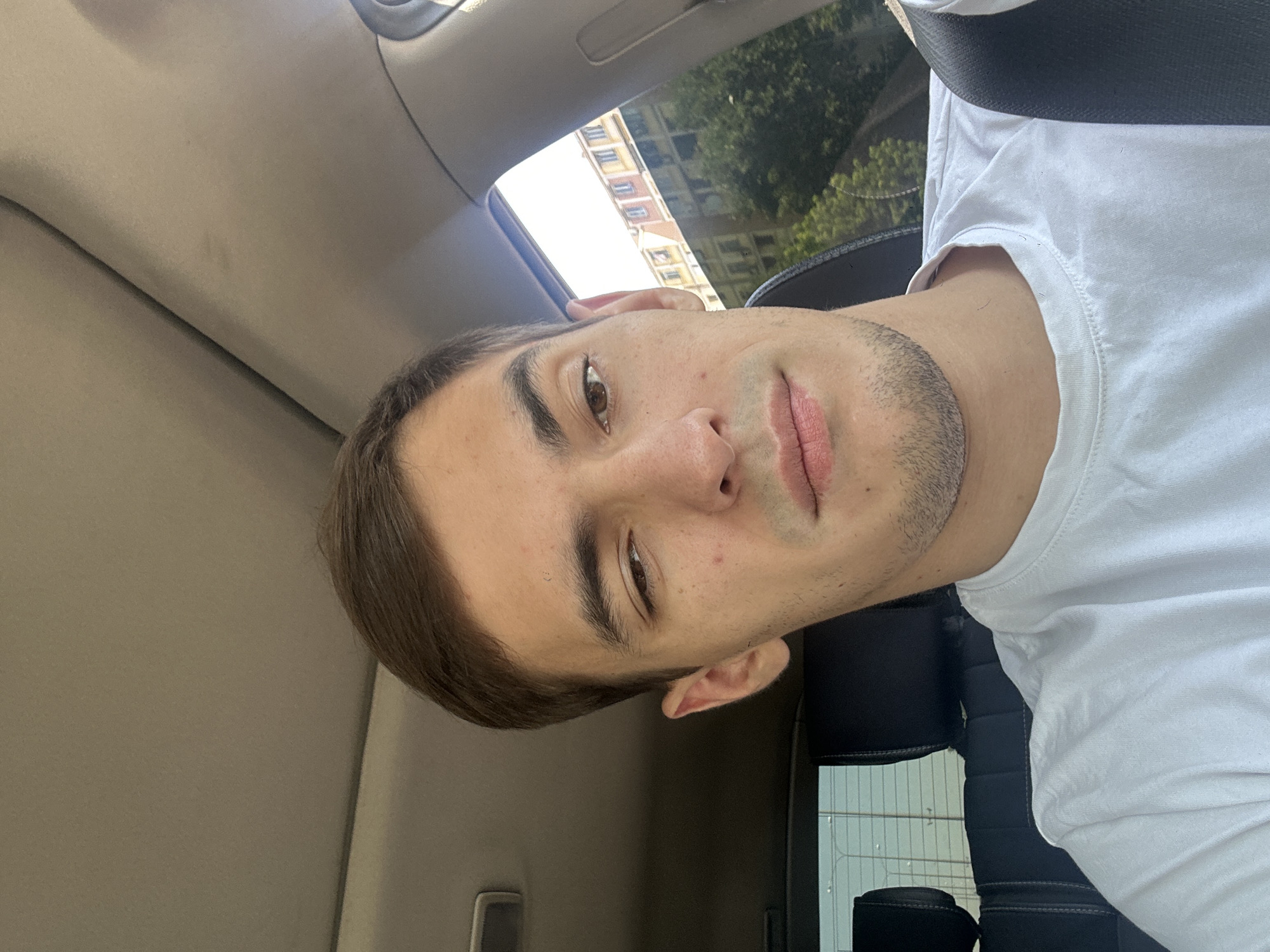 A close-up selfie of a young man with light brown hair and brown eyes is taken inside a car. He is wearing a white t-shirt and his left eye is partially closed. The background shows the car's interior, with the ceiling and dashboard visible, and through the window, buildings and trees are seen. The man appears to be in his early 20s. The presence of car interior details and city buildings in the background suggest an urban setting. Without further information or visual cues specific to Tashkent, Uzbekistan, it is not possible to definitively confirm the location.