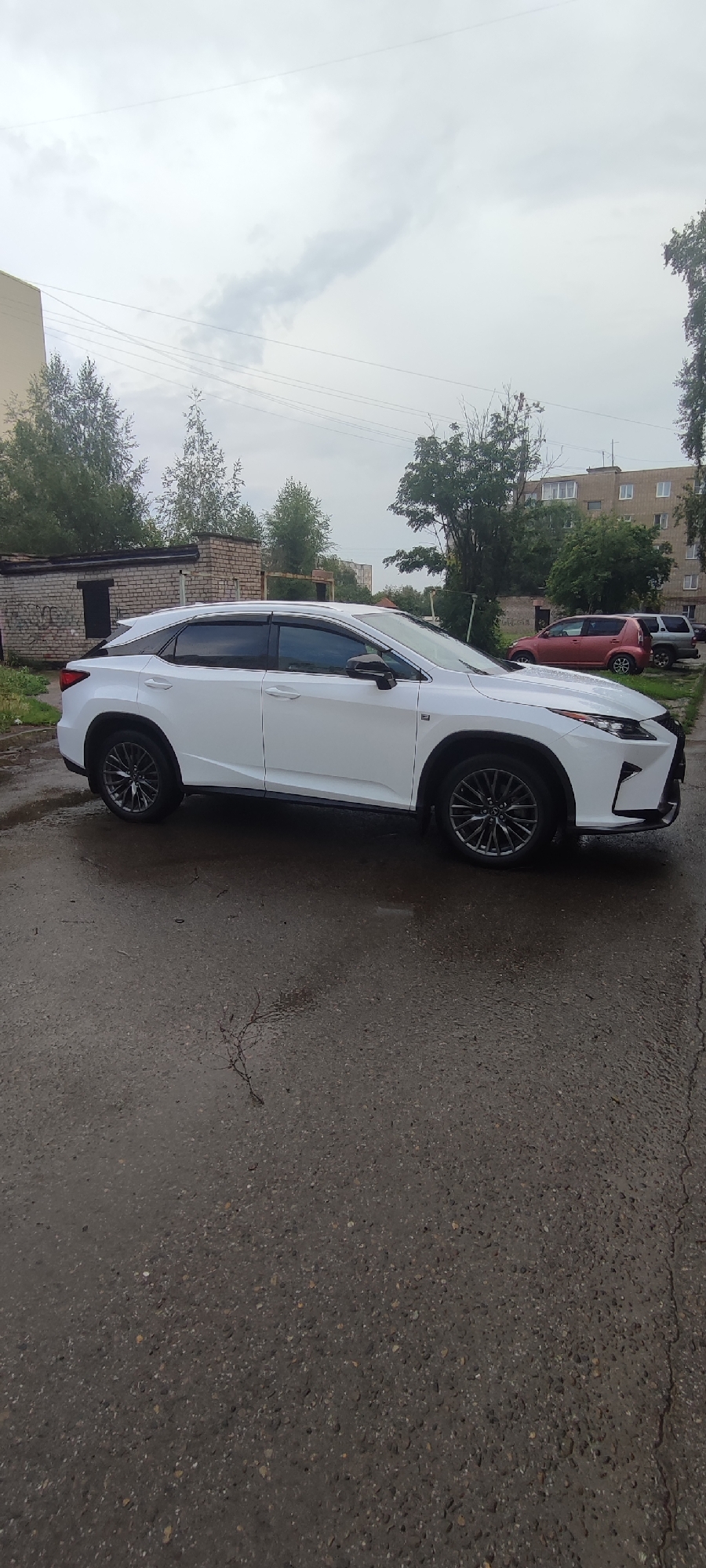 The image shows a white Lexus RX parked on a wet asphalt surface. The car is positioned in a side profile view, facing towards the left of the frame. Behind the car, there is a low brick building with a dark opening, possibly a garage or storage unit, and further in the background, a multi-story residential building with numerous windows. Trees and foliage are visible between the buildings and in the background. The sky is overcast with clouds. The scene appears to be a residential area in Novoaltaysk, Russia, likely on a day with recent precipitation. No people are present in the image.