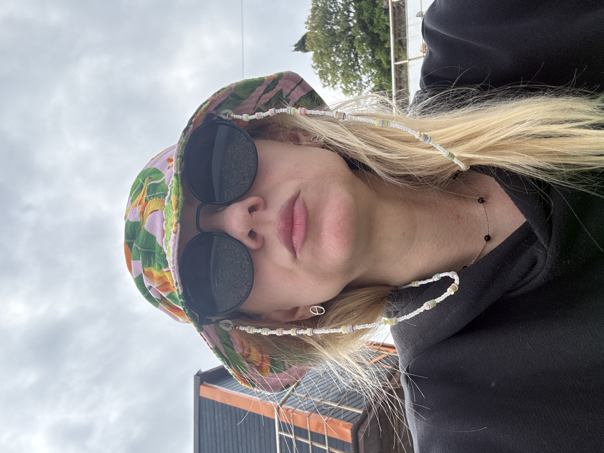 A fair-skinned individual with blonde hair is wearing round, black sunglasses, a patterned bucket hat, and a black sweatshirt. A beaded glasses chain is attached to the sunglasses. The background includes a cloudy sky, a tree, part of a building, and what appears to be a fence. The location is city of Unknown, Unknown.