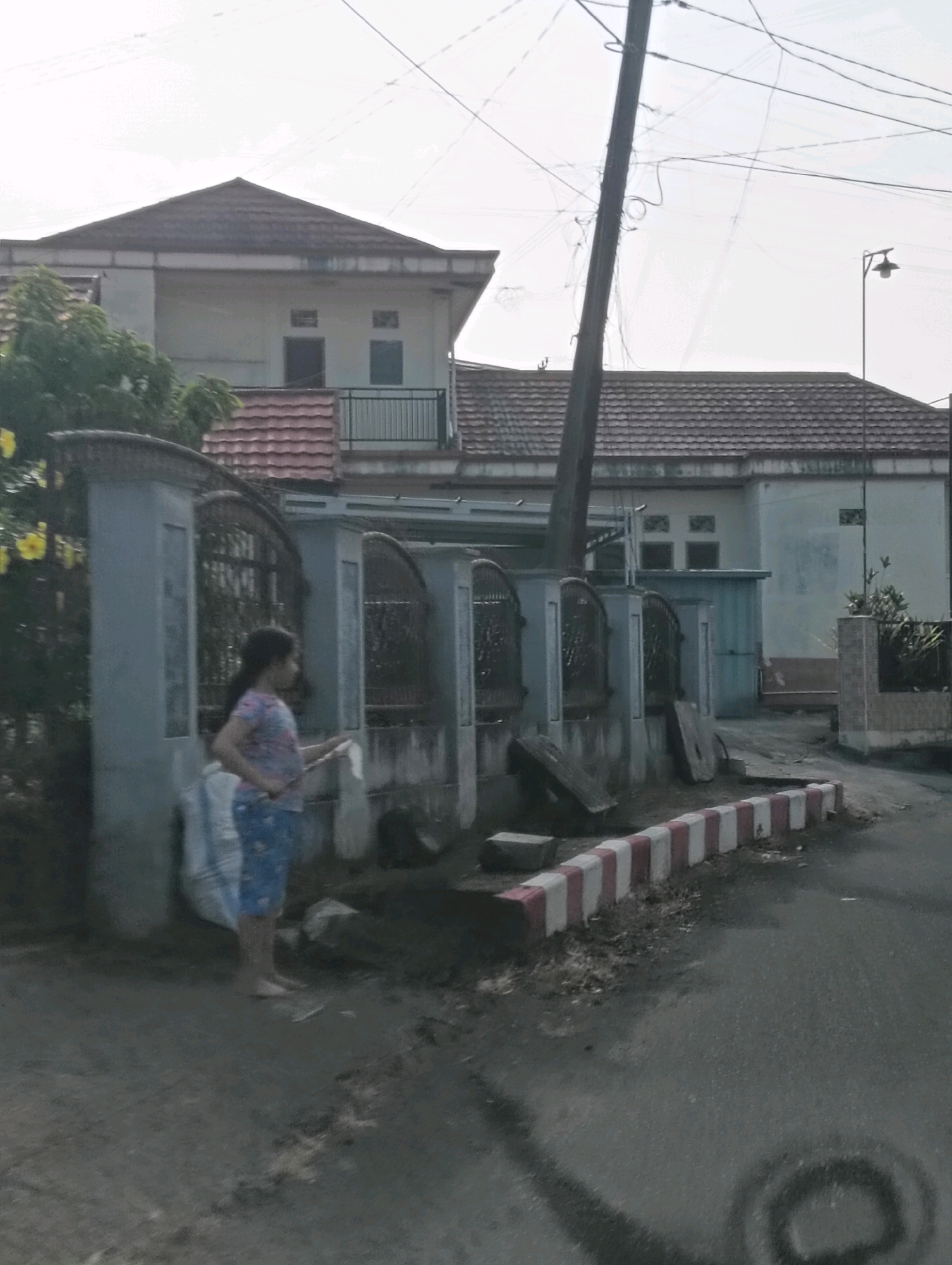 A young girl stands near a concrete fence with metal gates in front of a house in Tomohon, Indonesia. She is holding a large white plastic bag. The fence has decorative pillars and is partially covered with greenery. The house behind the fence has a tiled roof and windows on the upper floor. A telephone pole with many wires is visible leaning over the scene. The ground is a mix of dirt and asphalt, with some debris scattered around. A red and white striped curb runs along the edge of the road. The sky is overcast.
