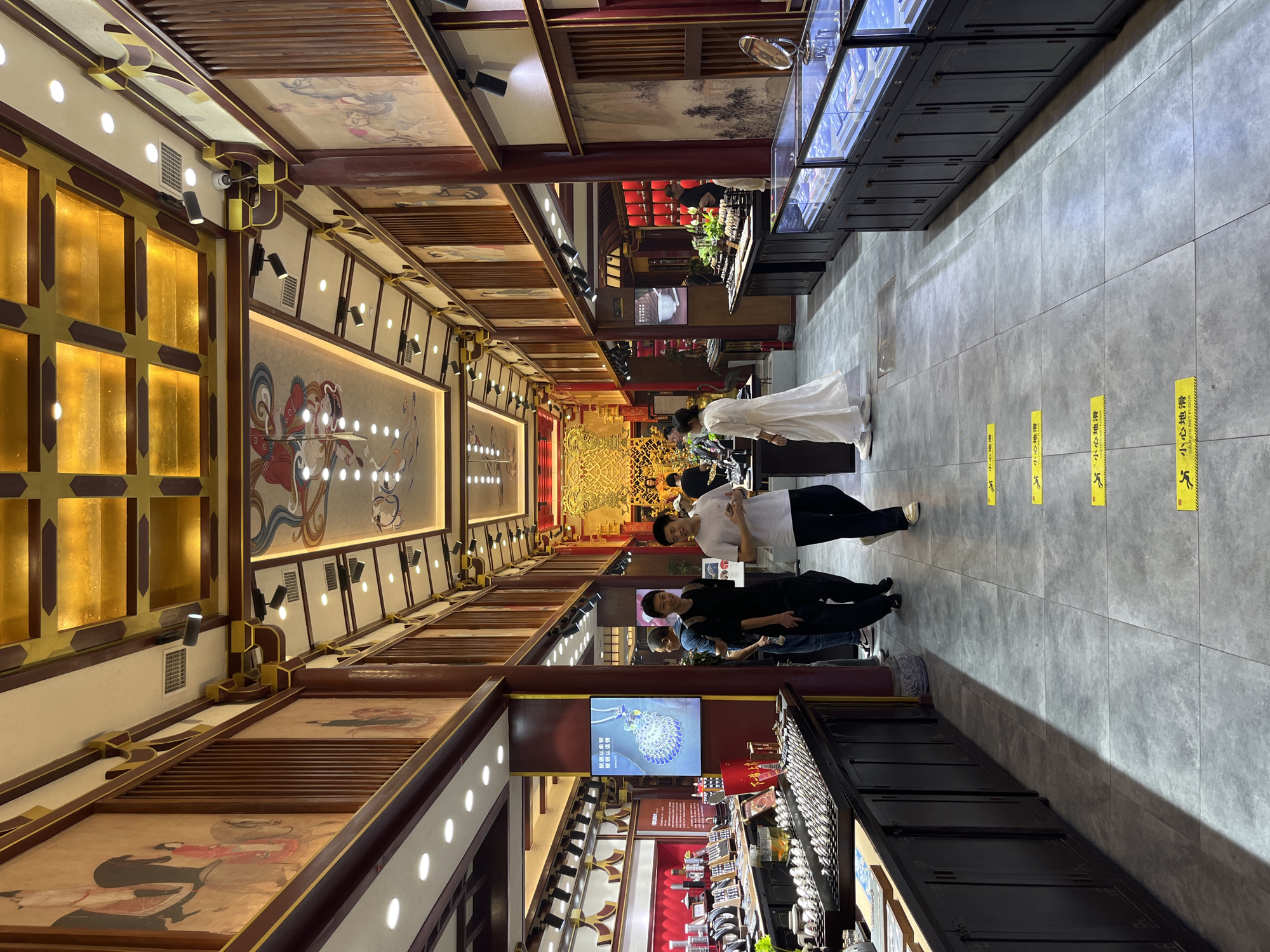 The image depicts the interior of a brightly lit, ornate jewelry store. The architecture features traditional Chinese elements, including red wooden pillars, intricately painted murals on the walls, and a golden, coffered ceiling with decorative artwork. Display cases filled with jewelry are arranged along the sides of the aisle. Several people are visible walking through the store. In the foreground, two men, one in a black t-shirt and the other in a white t-shirt, are walking away from the camera. Further down the aisle, a woman in a white dress is also walking. In the background, more display cases and ornate decorations, including a golden altar-like structure, are visible. Yellow caution signs with Chinese characters and an icon depicting a person slipping are placed on the floor, indicating a potential hazard.