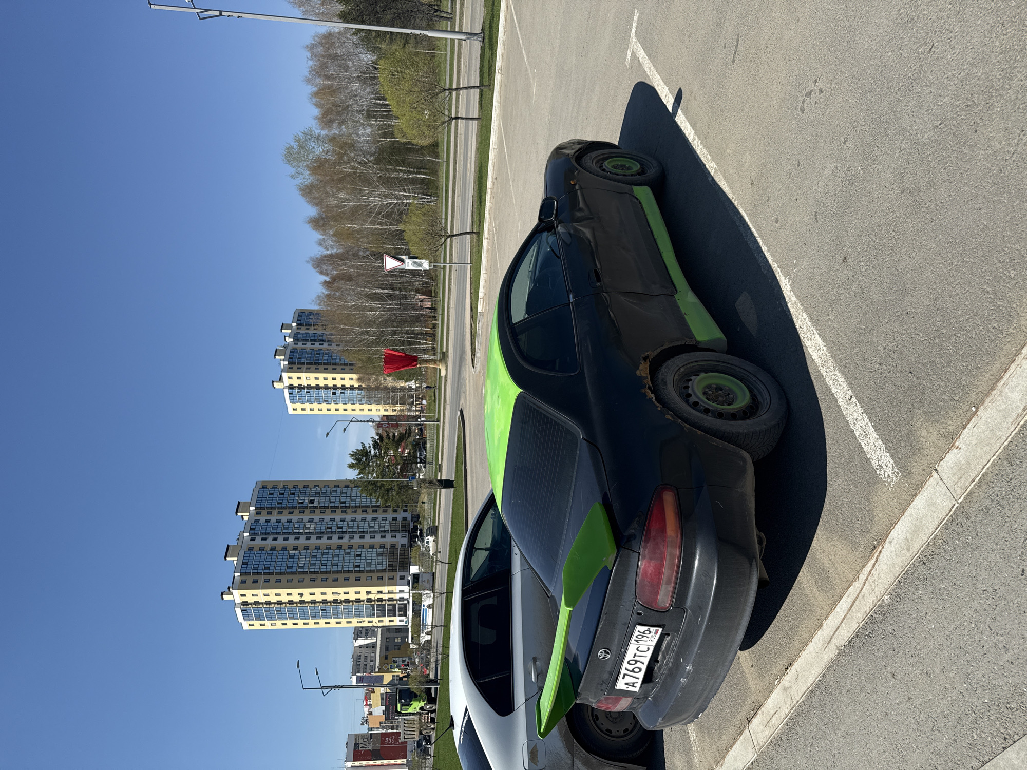 A parked, black and green, partially disassembled car with license plate A769TC196 is on paved asphalt in Tobolsk, Russia. Residential buildings are in the background, along with bare trees and a blue sky.