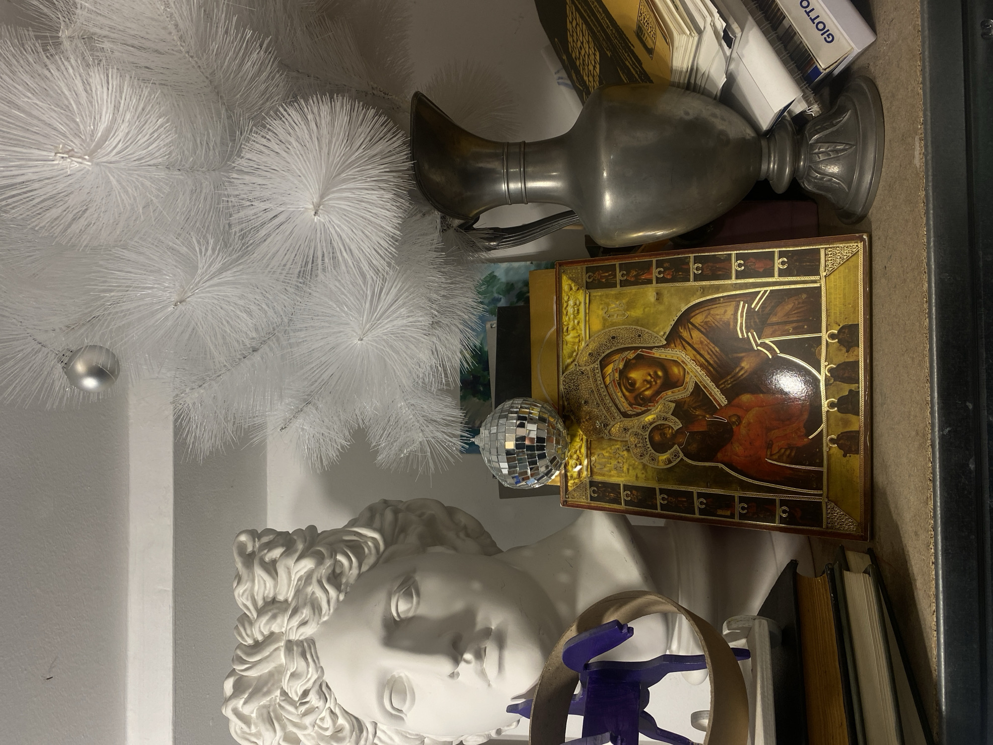 A still life displays various objects against a pale wall and dark background. A white, faux Christmas tree branch with a silver ornament hangs at the upper left. A white plaster bust of a human head lies in the lower-left corner. To the right of the tree and bust sits a stack of items including a silver pitcher, a disco ball, books, an icon of the Virgin Mary, and a box labeled "GIOTTO." In the foreground, in front of the bust, lies a tan strap with a dark blue object attached. The scene's location is Unknown, Unknown.