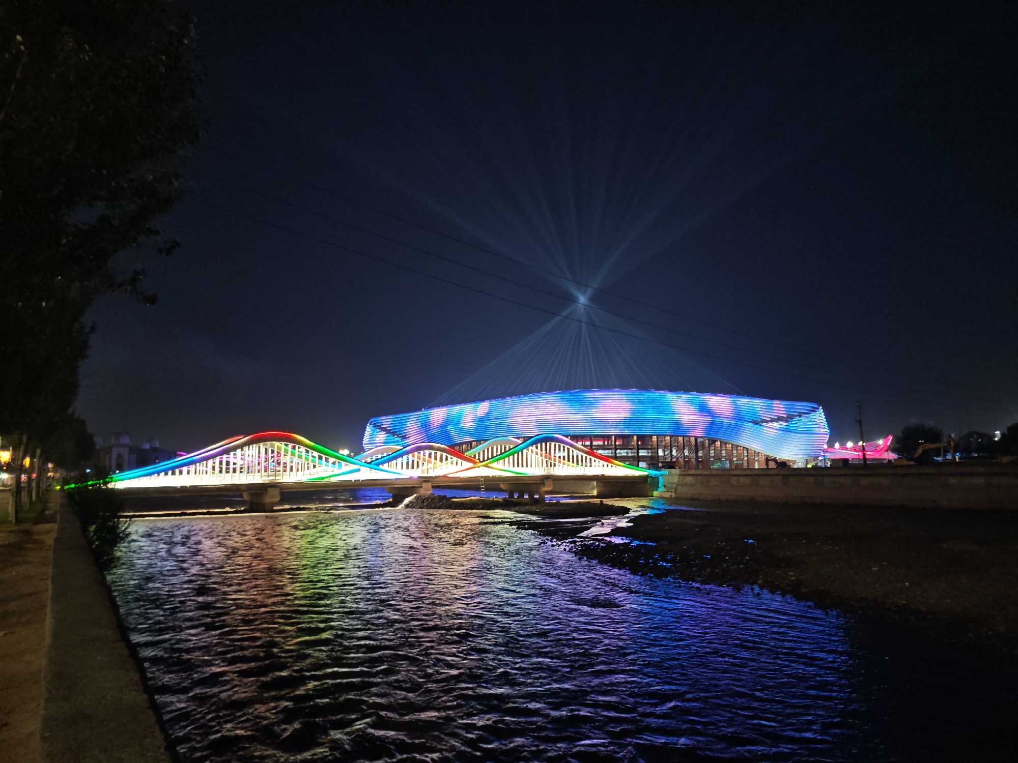 This is a nighttime photograph of a brightly lit bridge and building complex situated on a riverbank in Dushanbe, Tajikistan. The bridge spans across a dark body of water, with its arches illuminated by a vibrant rainbow of colors, including green, blue, red, and yellow. The water reflects these colors, creating a shimmering effect on its surface.
In the background, a large, modern building is also illuminated with colorful lights, predominantly blue and pink, in a wave-like pattern. Beams of white light emanate from this building into the dark night sky, creating a dramatic spotlight effect. Power lines are visible crossing the sky above the scene.
To the left of the image, a dark tree with sparse foliage stands near a paved walkway, with faint lights visible in the distance along the street. The riverbank itself appears to be made of concrete or stone in the foreground and transitions to a rocky, exposed area further back, particularly on the right side of the river. The overall scene depicts a well-lit urban landscape at night, highlighting architectural features with dynamic light displays. There are no clearly visible people or specific actions depicted in the image.