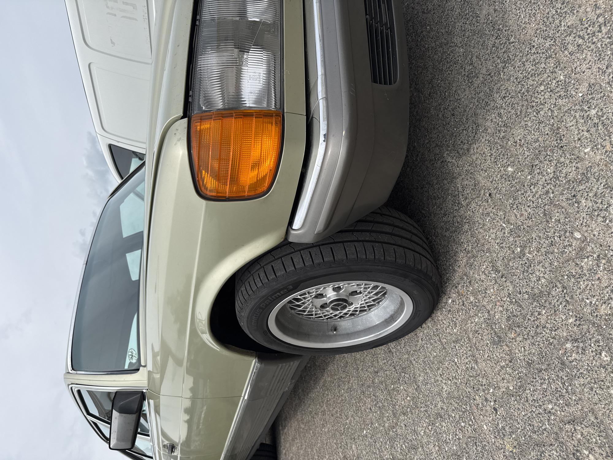 An outdoor, eye-level photograph, rotated clockwise, captures the front right quarter of a light green/beige classic car, likely a Mercedes-Benz W126 S-Class. The car is parked on a grey asphalt surface, characteristic of a street or parking lot. The overall lighting is diffuse, suggesting an overcast day.
The car's front right rectangular headlight features a clear lens and an integrated orange turn signal. Below the headlight, a grey bumper with a chrome strip shows some minor scuffs and wear. The front right wheel is fitted with a multi-spoke (BBS-style) alloy rim, featuring a silver polished lip and a Mercedes-Benz star in the center cap. The tire, a Hankook Ventus Prime 3, size 225/45 R17, is clearly visible.
Reflected in the car's hood and windshield is the side of a white van, possibly a Ford Transit, with faint, partially legible text such as "MA...STORE" on its side. The sky in the background is light grey and cloudy. Further back, the car's side profile reveals chrome trim around the windows and along the lower body. A black side mirror with a silver reflective surface is also visible. A small, round sticker can be seen on the lower part of the windshield. The scene is set in Lübeck, Germany.