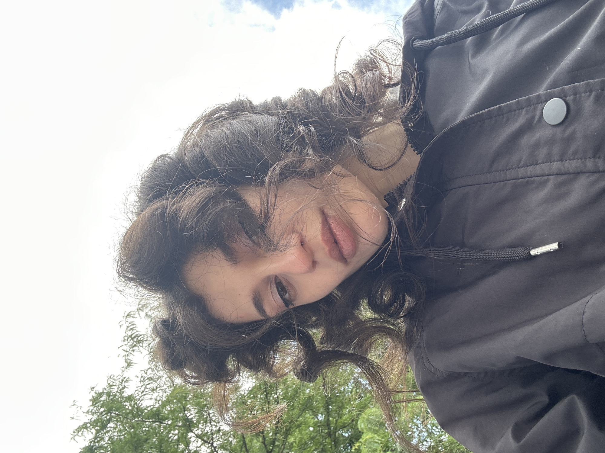 A young woman with curly brown hair, wearing a black jacket, poses for a selfie outdoors. Her hair is styled in large curls that frame her face and blow around her. She has subtle eye makeup and her lips are slightly parted. The background shows a sky with some clouds and green trees. The image is cropped from a low angle, with the woman's face and upper body taking up most of the frame. There are no distinguishing landmarks in the background that would indicate a specific location in Sofia, Bulgaria.