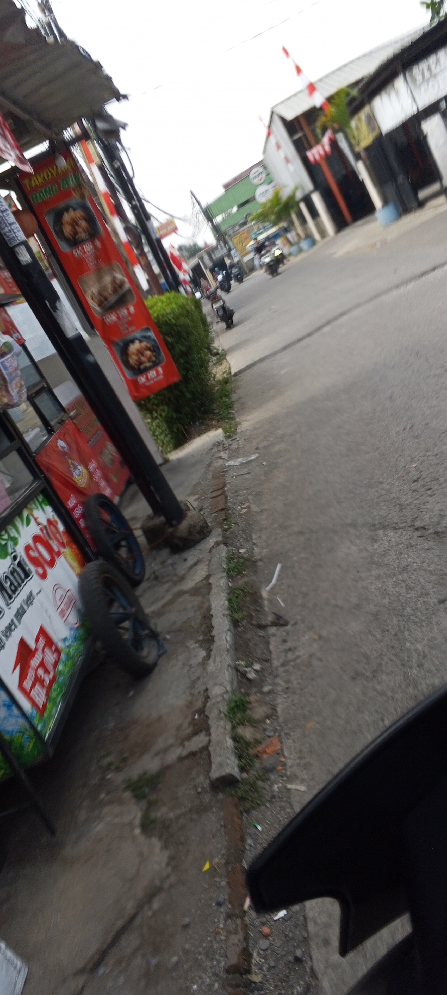 The image shows a street scene in Depok, Indonesia, likely during the daytime. The perspective is low, suggesting it was taken from a moving vehicle, possibly a motorcycle, looking forward and to the right. In the foreground on the left, there is a food cart or stall with a banner that reads "Nani SOLO" and other text in Indonesian. The banner features images of food, and price information is visible. Two wheels of the cart are clearly visible, one larger than the other.
To the right of the cart is a sidewalk with some vegetation and what appears to be debris. Beyond the sidewalk is a paved road with several motorcycles driving along it. In the mid-ground, buildings are visible along the street, some with signage. One building has a green facade and the word "VAPE" is discernable. Red and white Indonesian flags are displayed on poles and building eaves, indicating a festive or patriotic occasion. The sky is overcast. The overall impression is a typical street view in a busy Indonesian city.