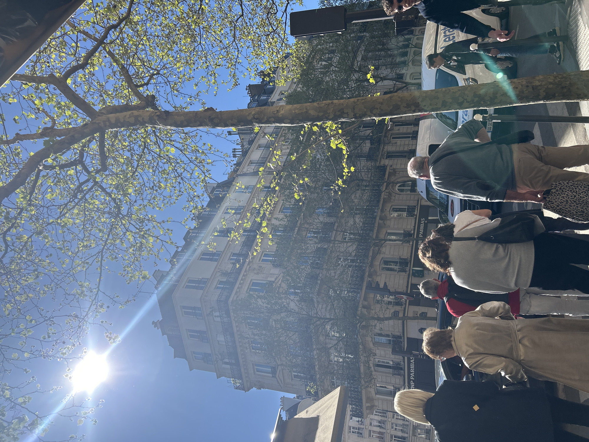 The image depicts a city street scene, likely in Paris, France, as a "BNP Paribas" sign is visible. A multi-story building with numerous windows and wrought iron balconies stands as a backdrop. Several people are walking on the sidewalk, including a man in a green shirt and khaki pants, a woman in a gray sweater and black pants, and others in various attire. The sun shines brightly in the sky, partially obscured by the branches of a tree with budding leaves. A white van and a man leaning on a street lamp are also visible.