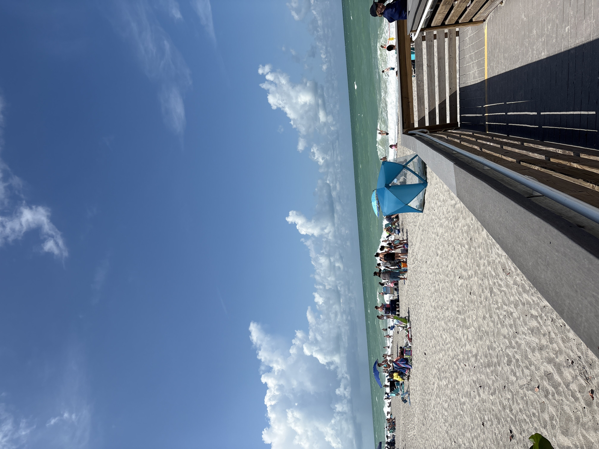 The image depicts a crowded beach scene on a sunny day. The sand is light in color, with numerous people scattered across it, many relaxing under umbrellas or in beach chairs. A turquoise ocean with moderate waves stretches across the horizon under a bright blue sky with scattered white clouds. To the right, a wooden boardwalk leads down to the beach, with railings and a yellow line marking a path. Several individuals are visible on the boardwalk and on the beach, some wading in the water or walking along the shoreline. A blue pop-up beach tent is set up on the sand. The overall impression is a vibrant and active recreational area.