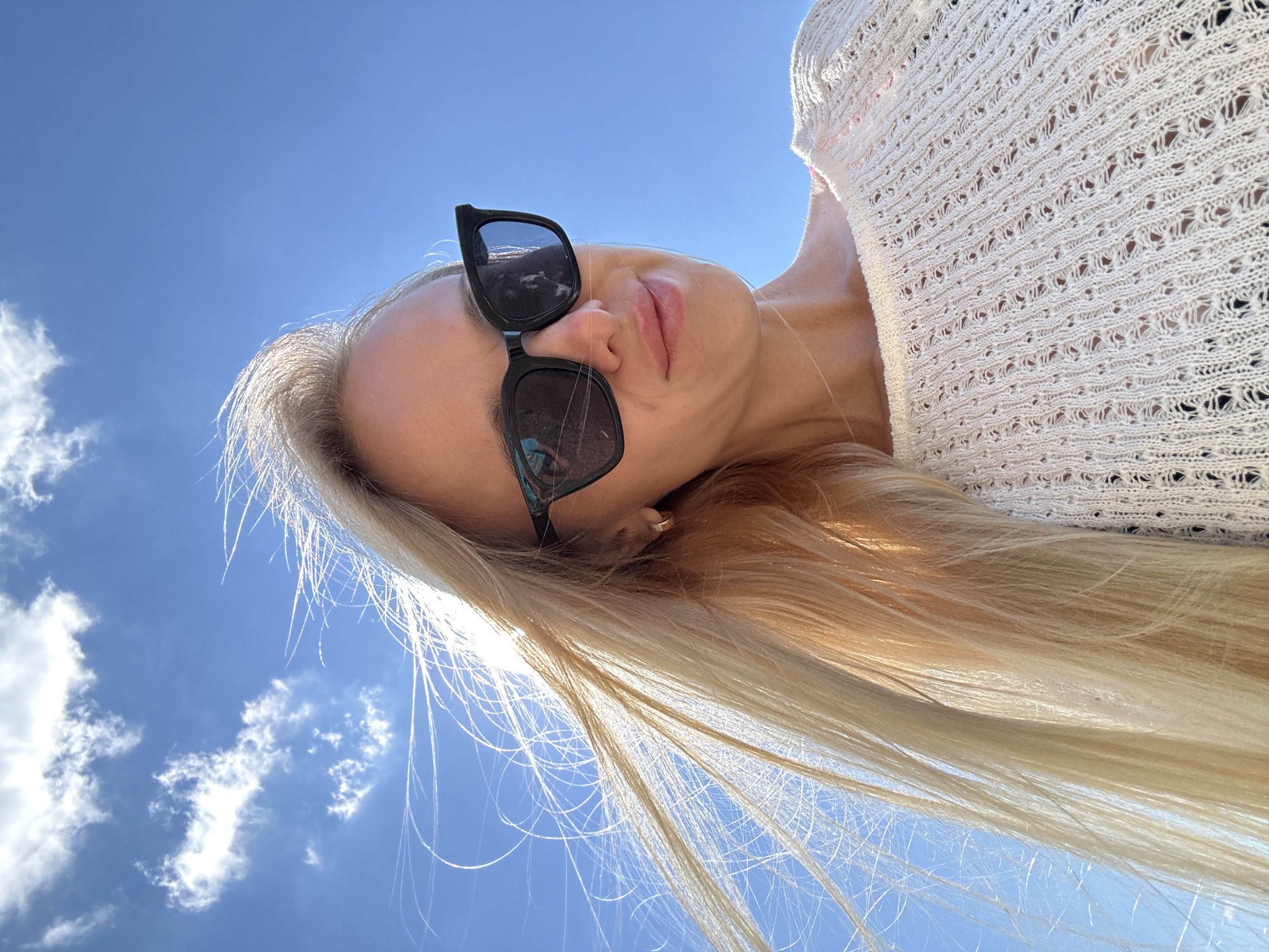 In Gornyi Erik, Russia, a young blonde woman with long, wind-swept hair and black sunglasses poses for a selfie against a clear blue sky with scattered white clouds. She is wearing a white, loosely knit sweater with an openwork pattern. Her hair is illuminated by sunlight, creating a bright halo effect. The perspective is a low-angle shot, emphasizing the vastness of the sky above her.