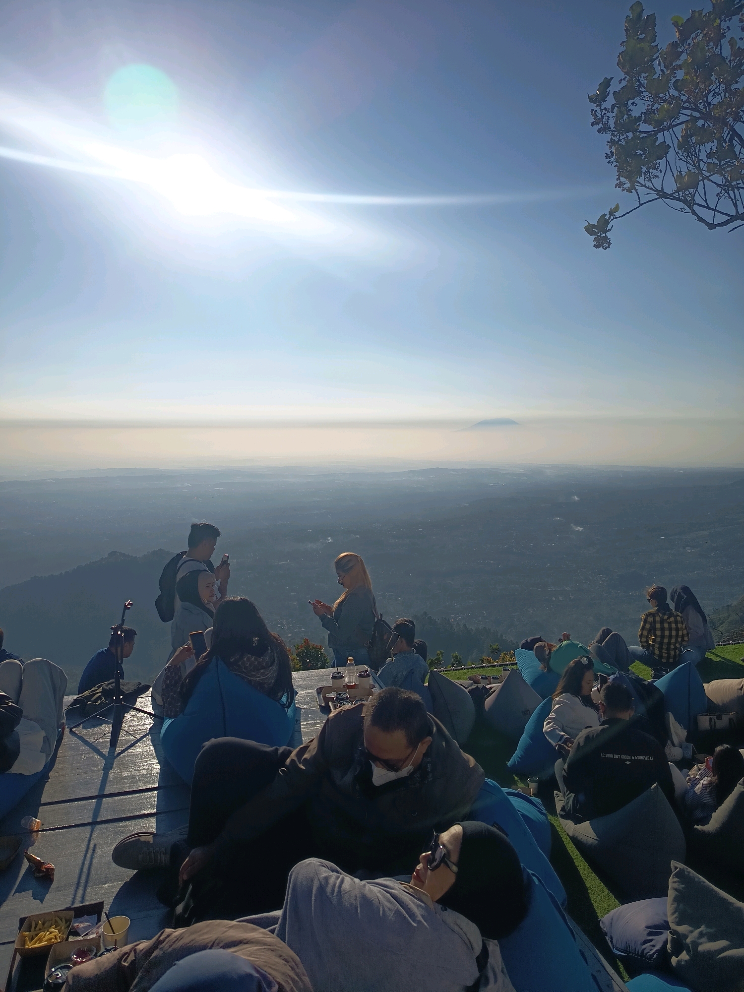 The image depicts an outdoor gathering of people, likely at a viewpoint overlooking a vast valley. The scene is bathed in bright sunlight, with a lens flare from the sun prominently visible in the upper left quadrant. People are seated and lounging on beanbag chairs, with some gathered on a wooden deck and others on a grassy area. In the background, a hazy landscape unfolds, featuring rolling hills and a distant mountain peak. Several individuals are looking at their phones, while others are engaged in conversation or simply enjoying the view. The atmosphere appears relaxed and informal. There are a few food and drink items visible on a table in the foreground, including a box of french fries. The overall impression is that of a scenic recreational spot in Keditan, Indonesia, likely a popular destination for enjoying the natural landscape.