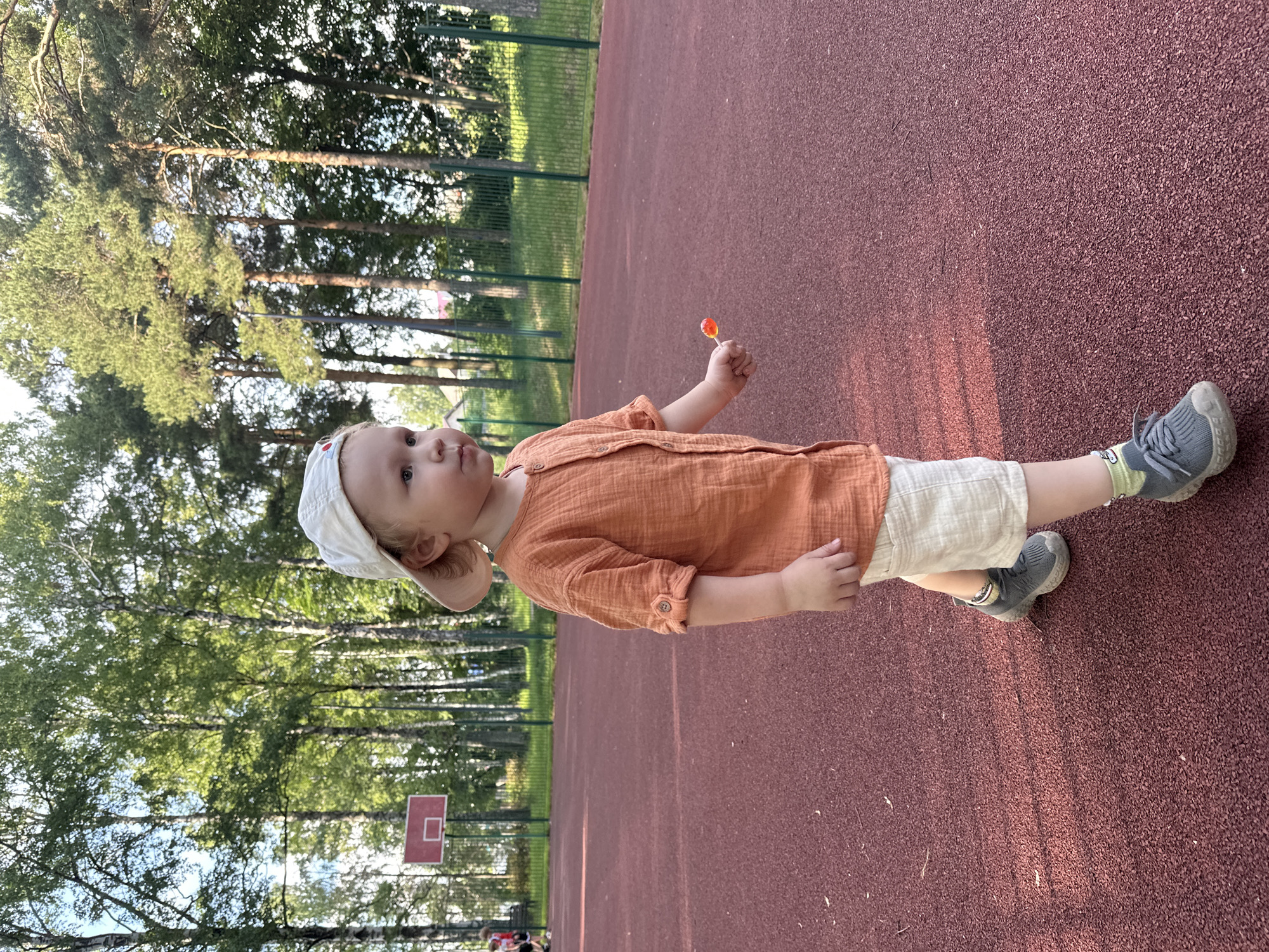 A young child wearing a white baseball cap, an orange gauze shirt, and light-colored shorts stands on a red outdoor court, likely a basketball court given the presence of a backboard and hoop in the background. The child is holding a lollipop in their right hand. The background consists of tall trees, a green metal fence, and a building. The scene appears to be in Votkinsk, Russia, based on the provided location information.