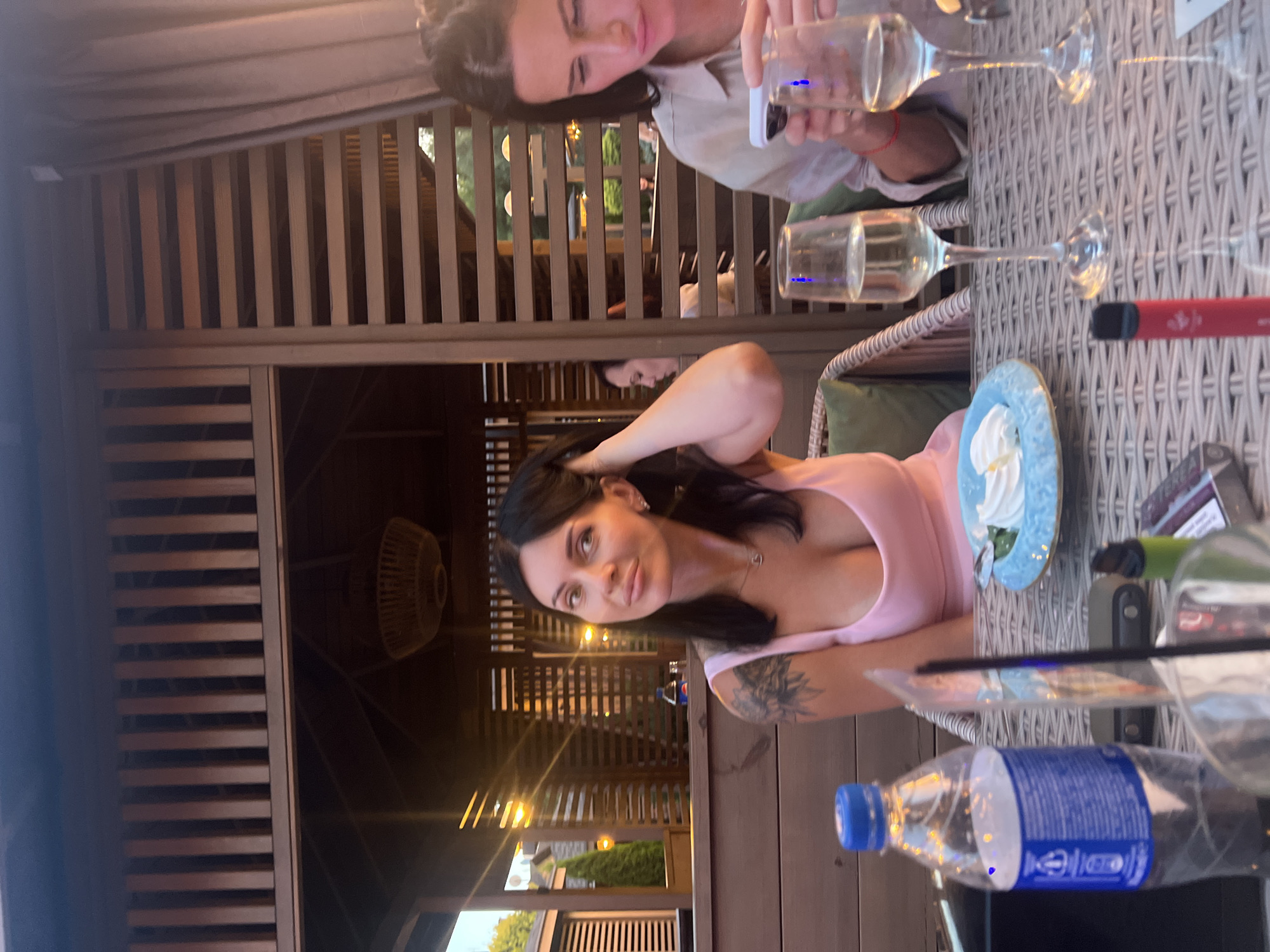 In an outdoor dining area, possibly a restaurant or patio, two women are seated at a table. The woman on the left, with dark hair and a pink dress, is looking towards the camera with her hand touching her hair. A blue plastic bottle of water and a plate with dessert are in front of her. The woman on the right, with dark hair and a light-colored shirt, is looking down at her phone and holding a white object. Two wine glasses filled with a pale yellow liquid are on the table between them. The background consists of wooden structures, possibly a pergola or gazebo, with hanging lights, and greenery visible through the gaps.