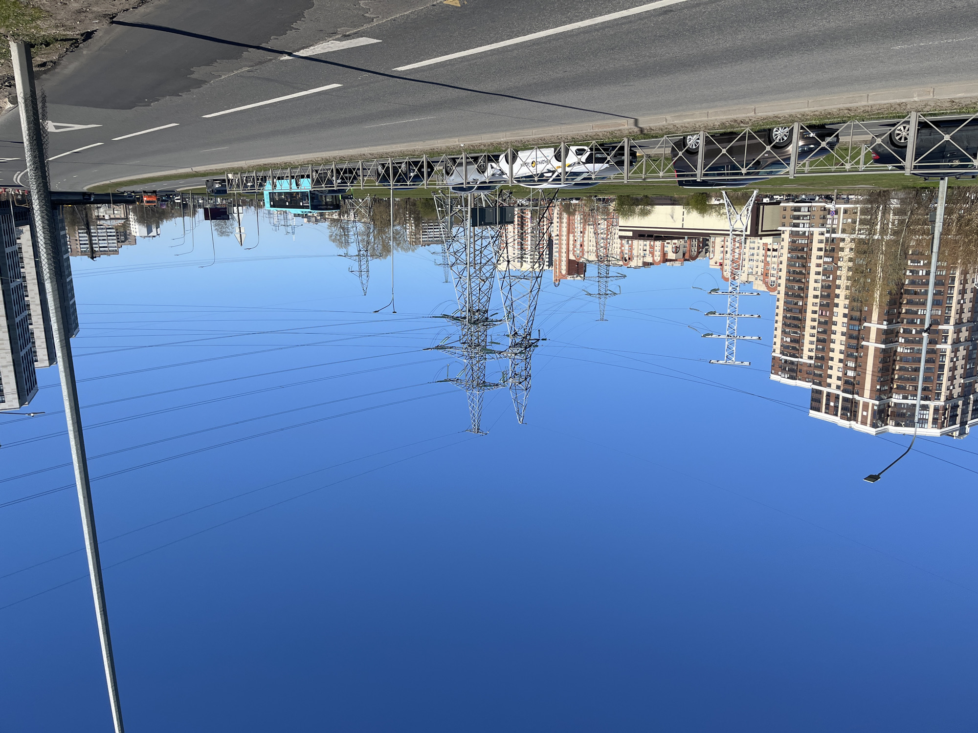 The image presents a bright, clear daytime view of an urban landscape, likely in Saint Petersburg, Russia. A wide, multi-lane asphalt road with distinct white lane markings spans the lower part of the frame. Several vehicles are visible on the road, including a light blue bus and various cars. A white car with the "Делимобиль" (Delimobil) car-sharing service branding and logo is prominently seen on the right, behind a metal pedestrian barrier.
The sky, occupying the majority of the upper frame, is a vibrant, cloudless blue. Dominating the central and upper-mid sections are multiple large, metallic lattice power transmission towers (pylons), from which numerous power lines stretch across the sky, running diagonally across the scene.
In the background, on both sides of the road, stand multi-story residential buildings, some featuring warm orange-brown facades and others lighter hues. Patches of green grass and sparse trees are visible along the road's edge, between the street and the buildings. A tall, grey concrete or metal pole extends upwards from the bottom-left corner of the image, while another street light fixture is visible suspended over the road on the upper right. The scene depicts a typical, busy urban environment under sunny conditions.