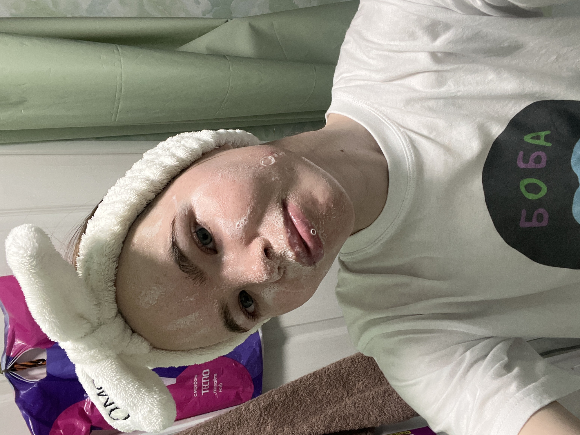 A person wearing a white t-shirt with "BOBA" written on it and a bunny ear headband is taking a selfie. The person's face is covered in a white substance. A purple package labeled "Tecno" is partially visible on the left side of the frame, next to a brown towel. Behind the person is a light green patterned surface. The scene appears to be indoors, and the packaging indicates the location might be Chaikovsky, Russia.