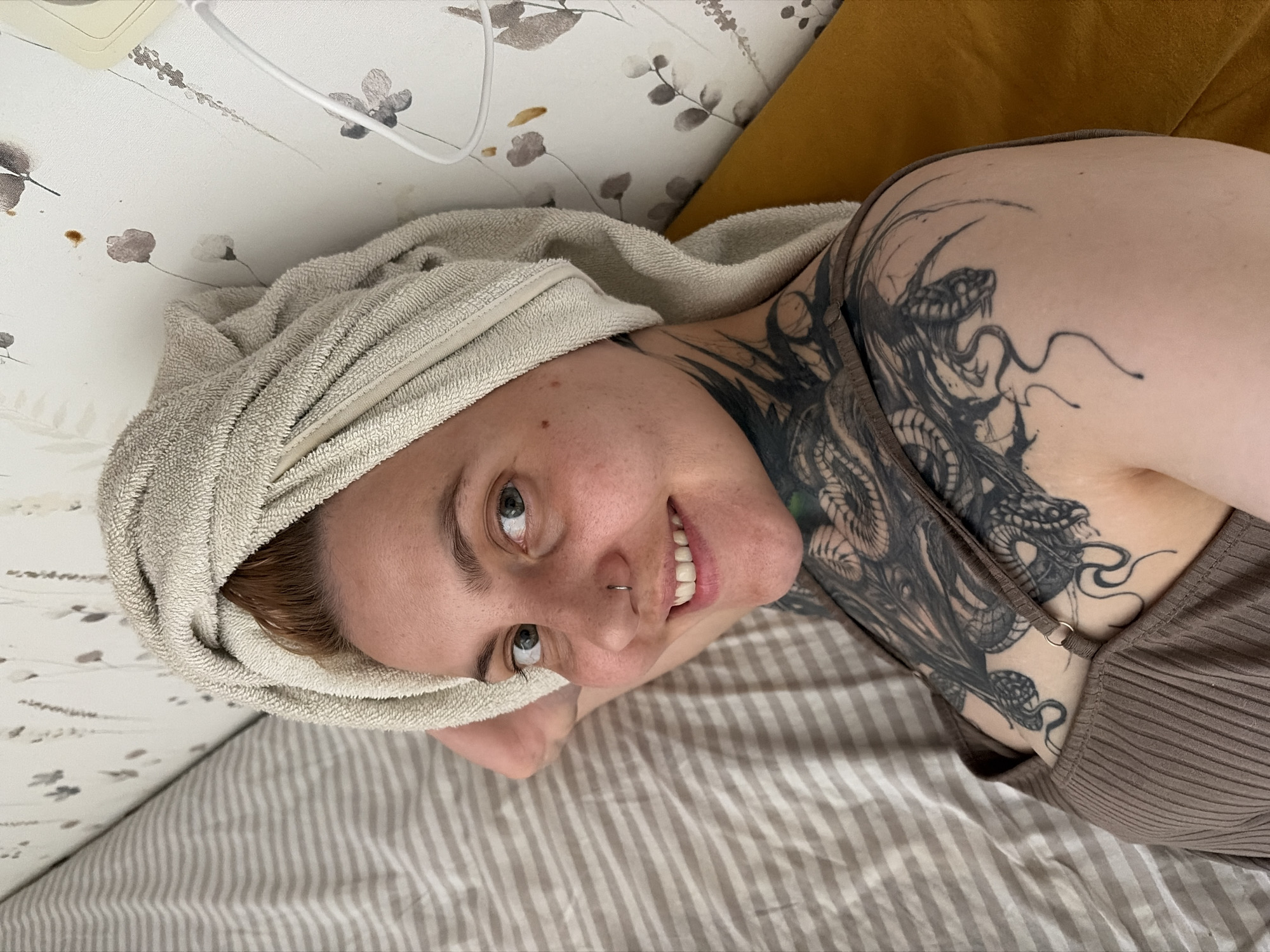 A woman lies on a striped bed in what appears to be a bedroom. She has a towel wrapped around her head, indicating she has recently showered. The woman has a snake tattoo on her chest and shoulder. She is smiling, and there is a nose ring on her left nostril. The background includes a headboard with a floral pattern and a mustard-yellow pillow. The scene seems domestic and personal. There is no information about the city of Chaikovsky, Russia.